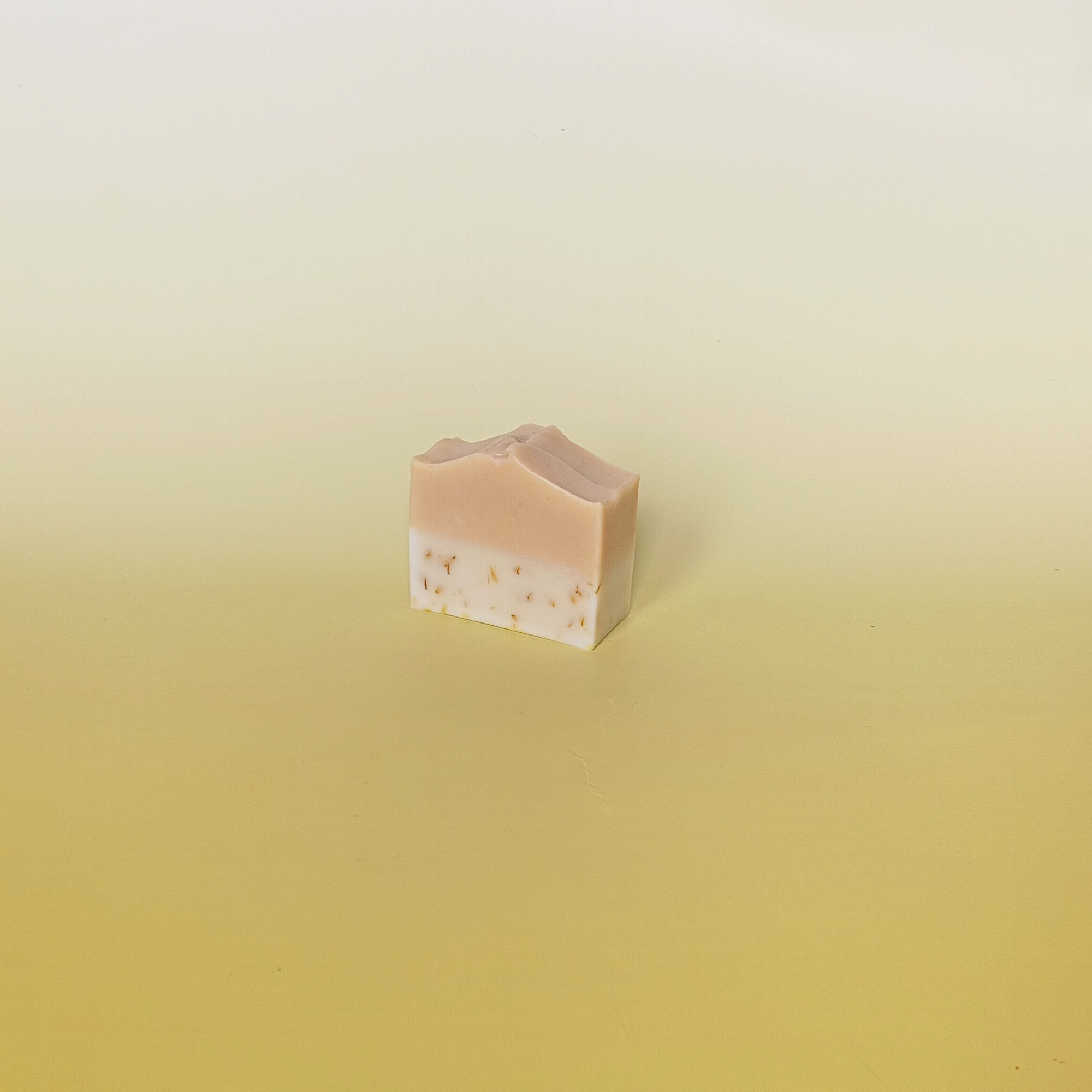 Handmade two-tone natural soap bar with soft blush and cream layers, displayed on a pale yellow gradient background, showcasing a minimal and gentle aesthetic.
