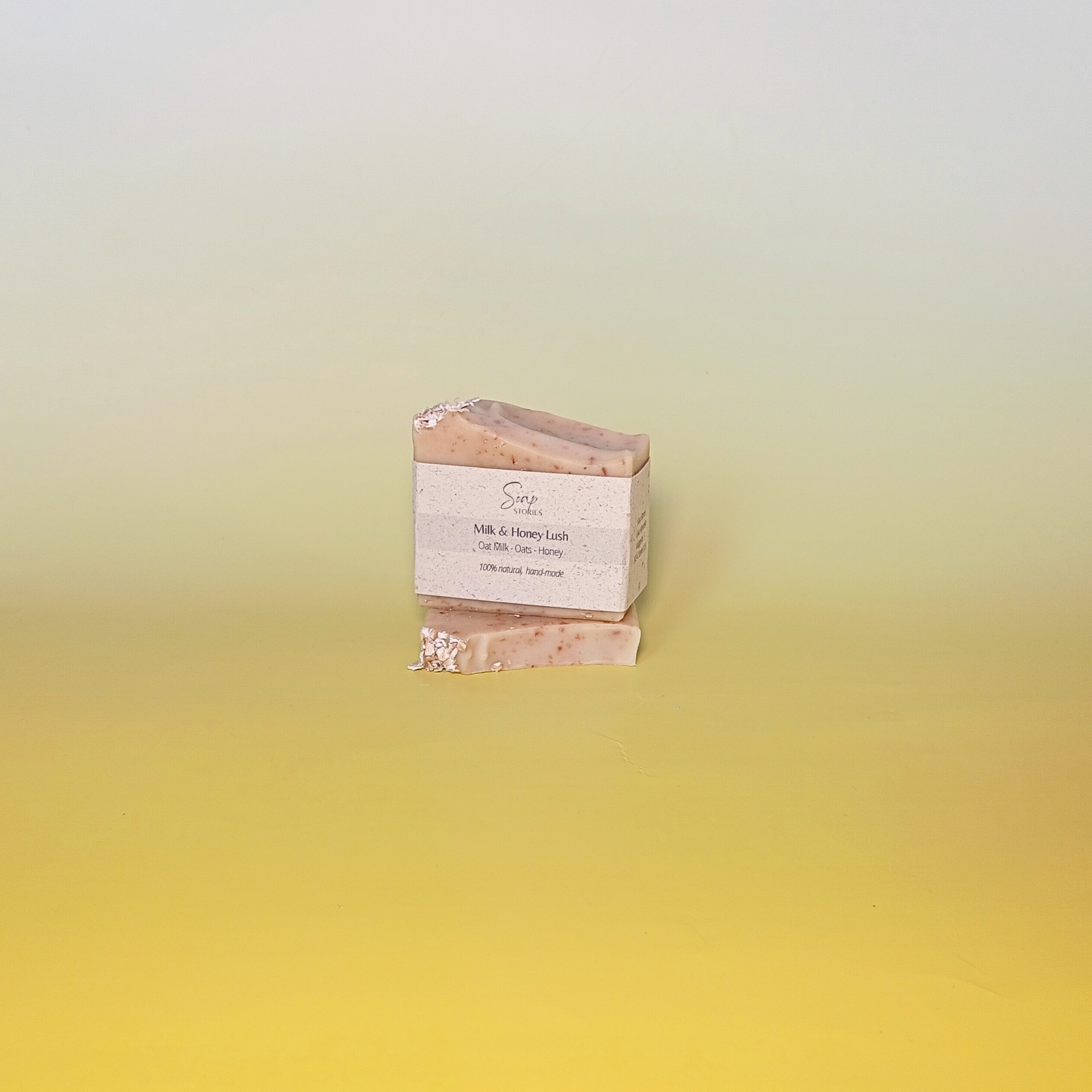 Handmade natural soap bar with a soft beige tone and visible oats, unscented and crafted with milk, honey, and oats, displayed on a pale yellow gradient background wrapped in grass paper sleeve