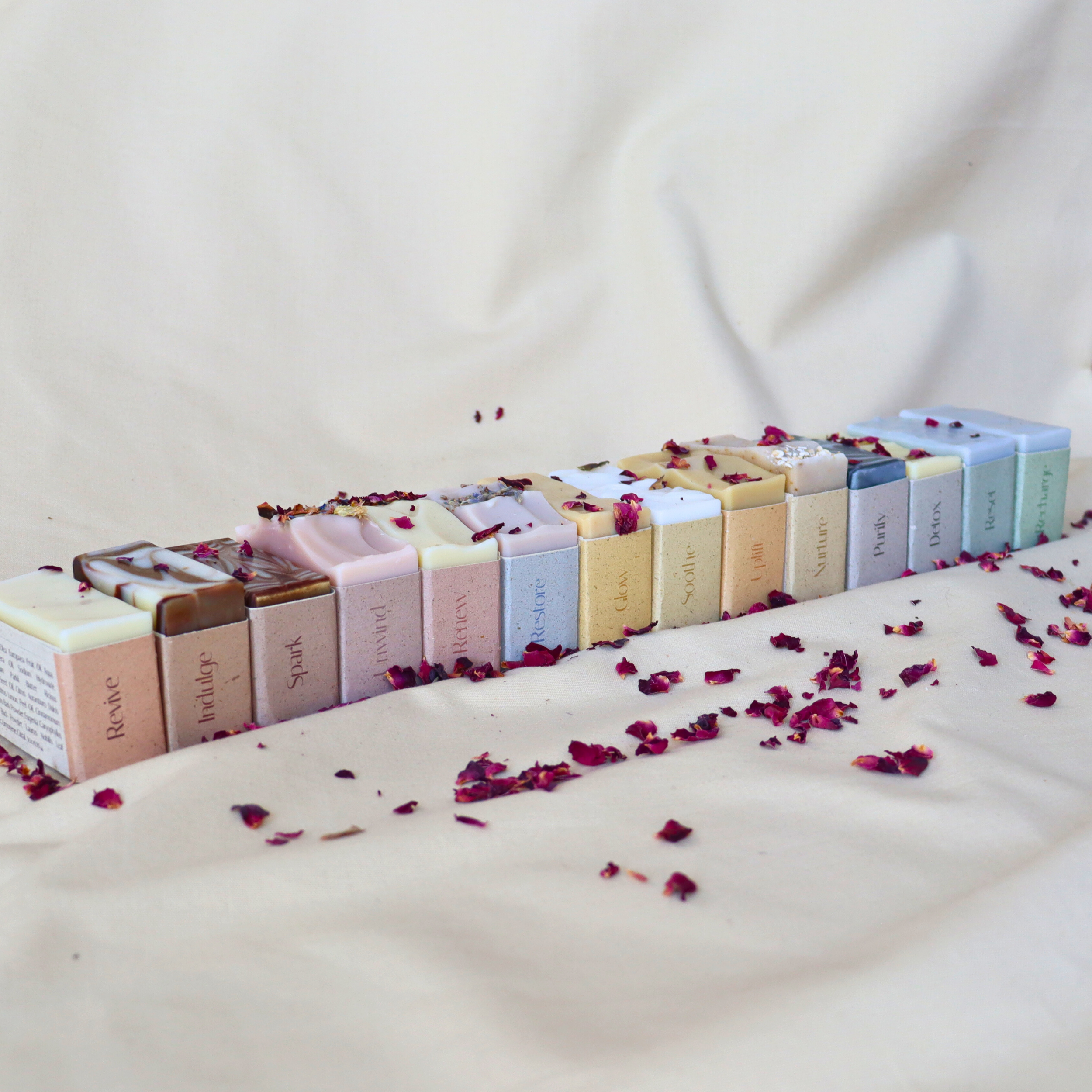 Colorful handmade soap collection arranged in a straight line with rose petals scattered on neutral fabric.
