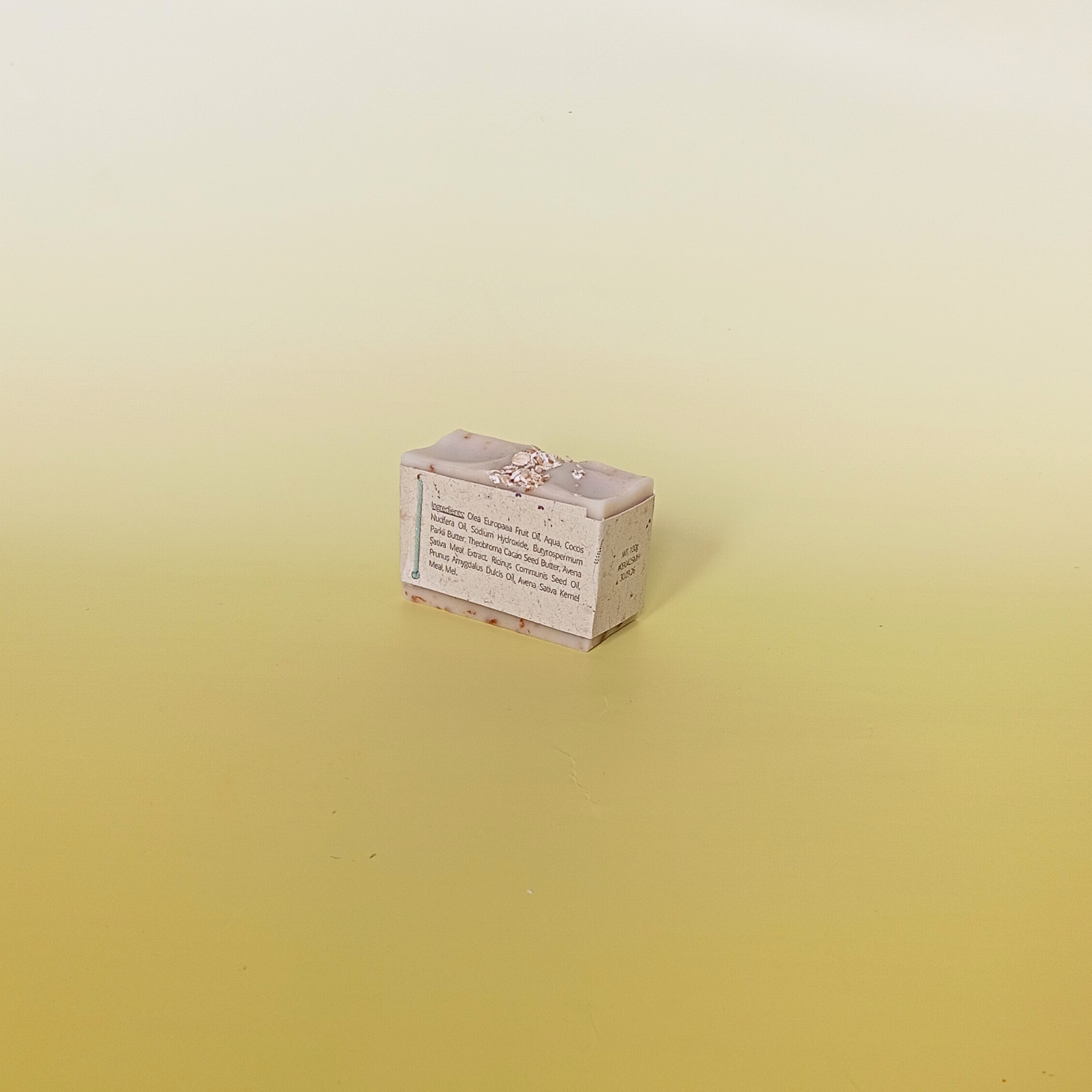 Handmade natural soap bar with a soft beige tone and visible oats, unscented and crafted with milk, honey, and oats, displayed on a pale yellow gradient background wrapped in a grass paper sleeve