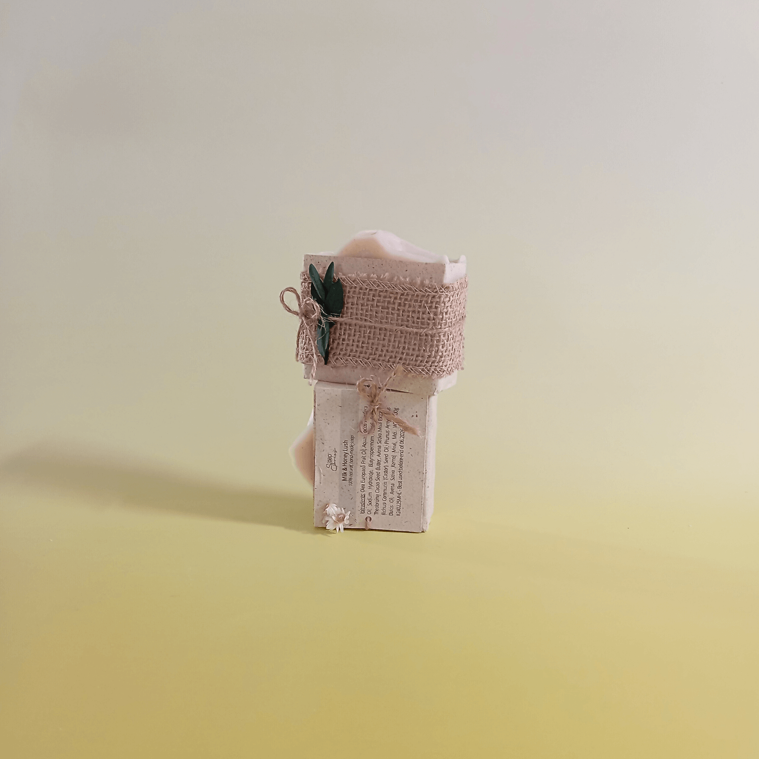 Natural soap bars wrapped in rustic burlap and twine with green botanical accent, displayed vertically with visible ingredients label on a soft yellow background.