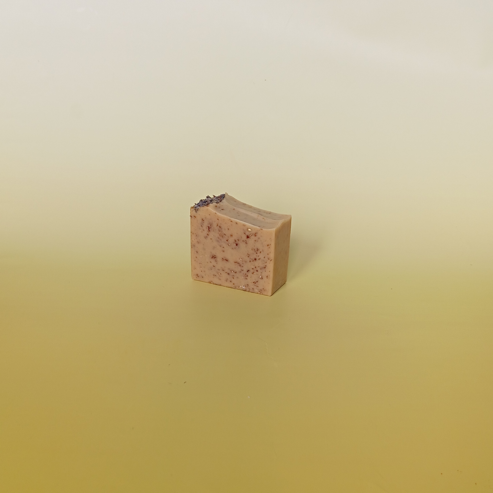 Handmade natural soap bar with a soft beige tone, featuring visible oats and topped with lavender buds, scented with milk, honey, and lavender, displayed on a soft yellow gradient background.