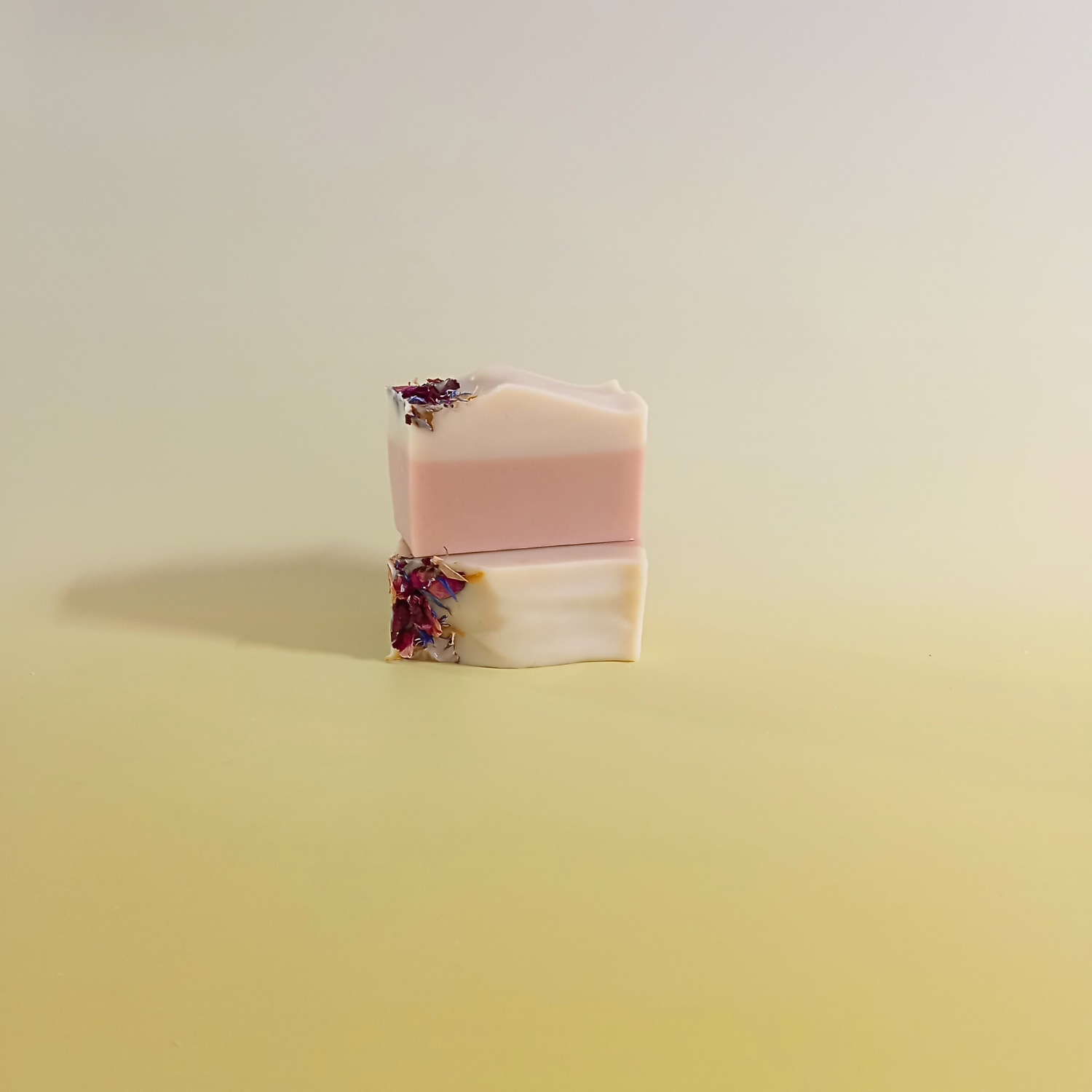 Two handmade natural soap bars in soft blush and cream tones, topped with dried flower petals and stacked on a pale yellow background.