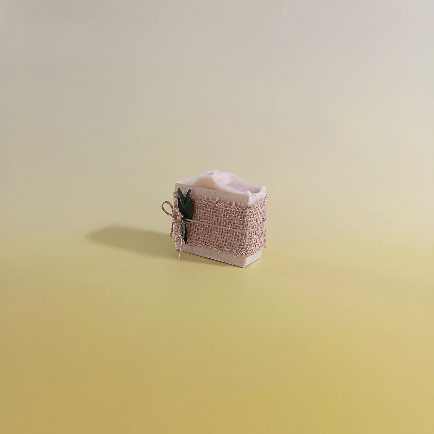 Natural soap bar wrapped in rustic burlap and tied with twine, accented with green botanical detail, displayed on a soft yellow gradient background for a natural and minimal look.