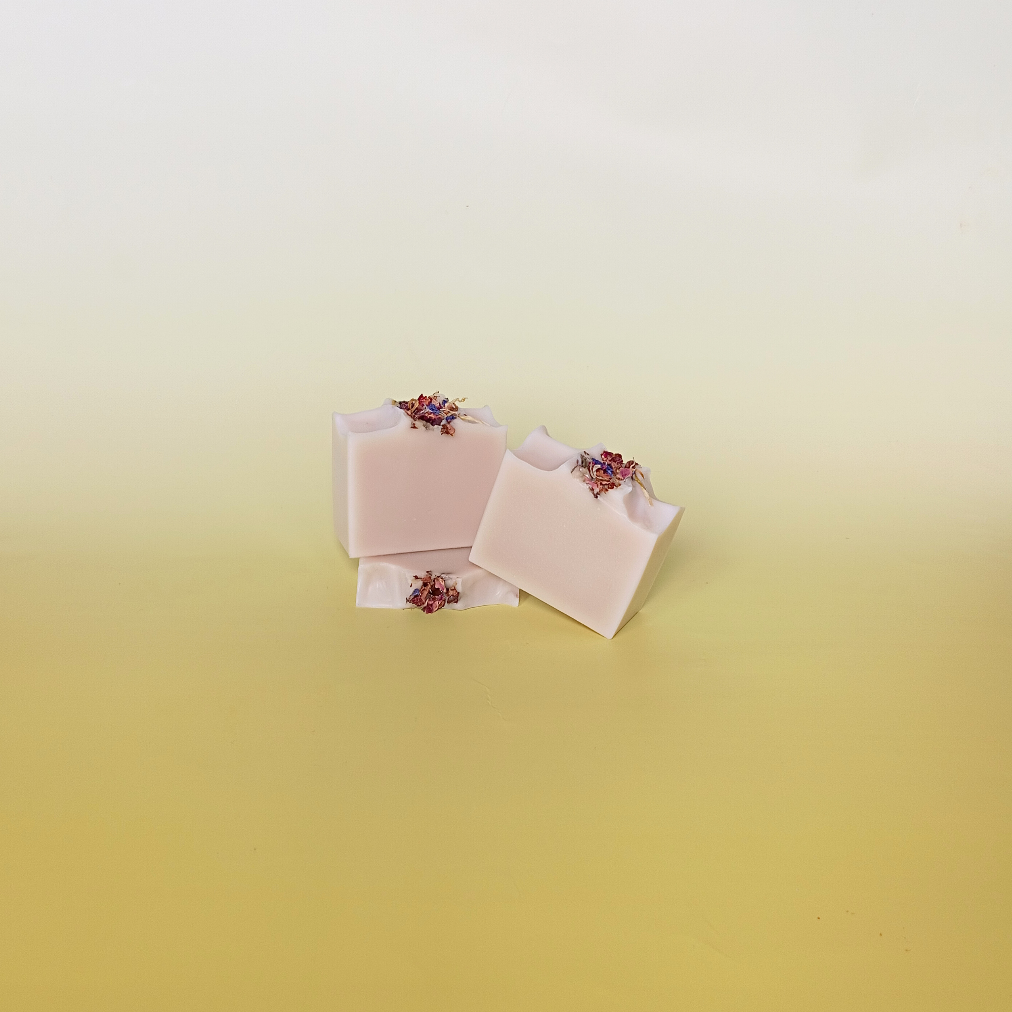 Three blush pink handmade soap bars with scattered dried petals, artistically stacked against a soft yellow gradient background, showcasing natural and minimal design.