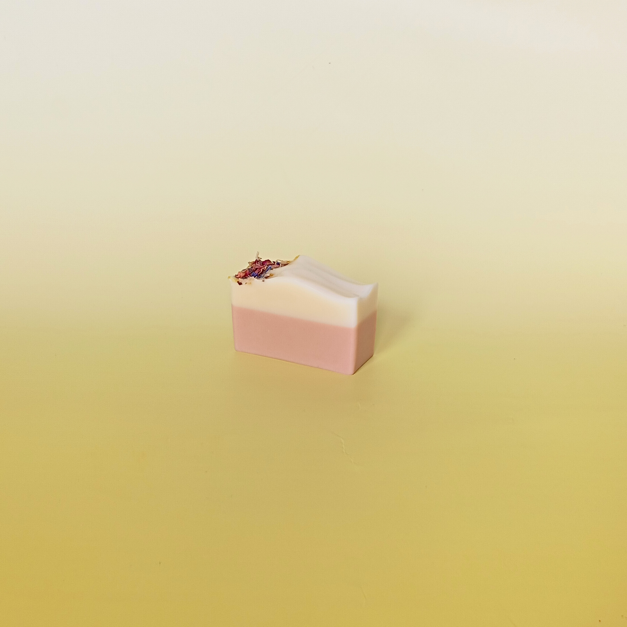 Handmade natural soap bar with soft pink and cream layers, crafted with kaolin and illite clays, scented with geranium and patchouli essential oils, displayed on a pale yellow gradient background.