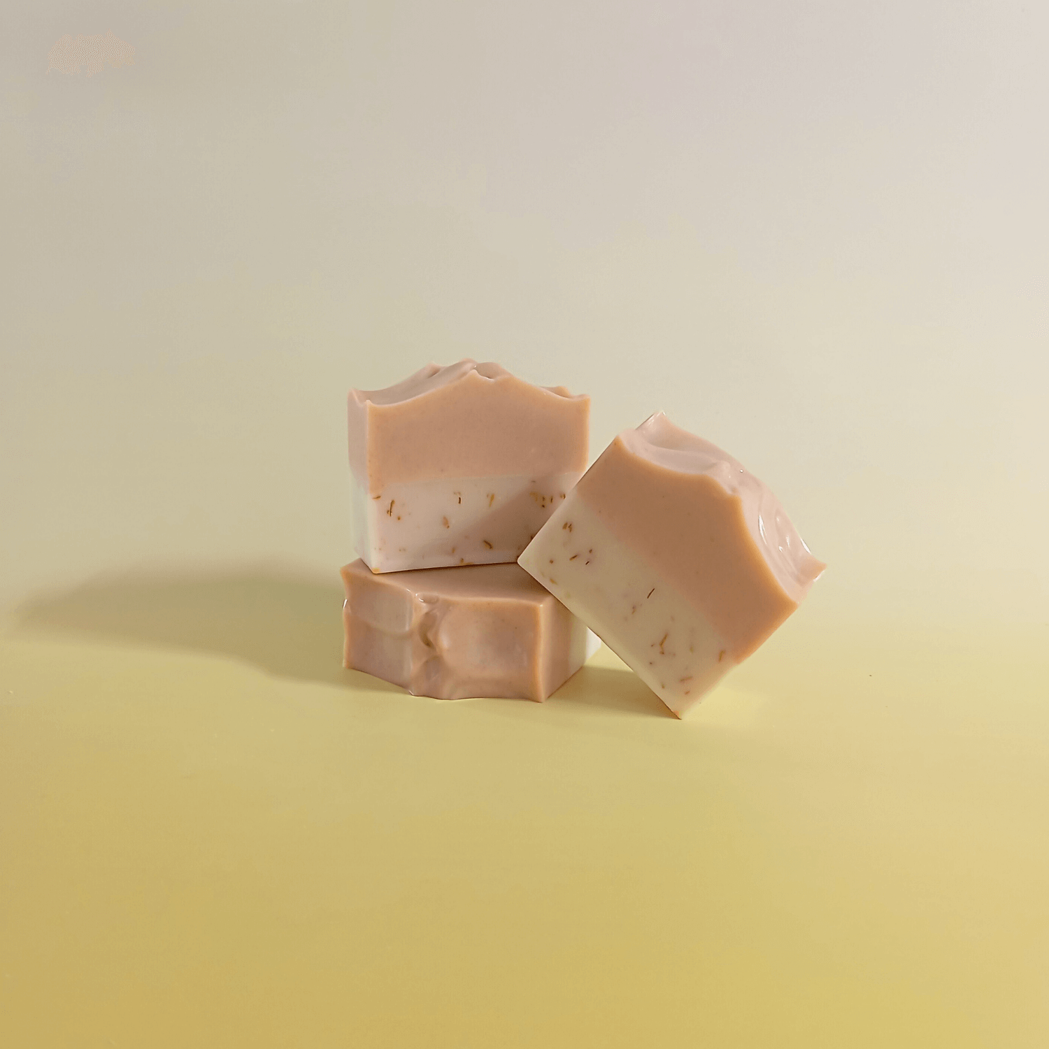 Three handmade two-tone natural soap bars with blush and cream layers, artistically stacked on a pale yellow gradient background, reflecting a soft and minimal design.
