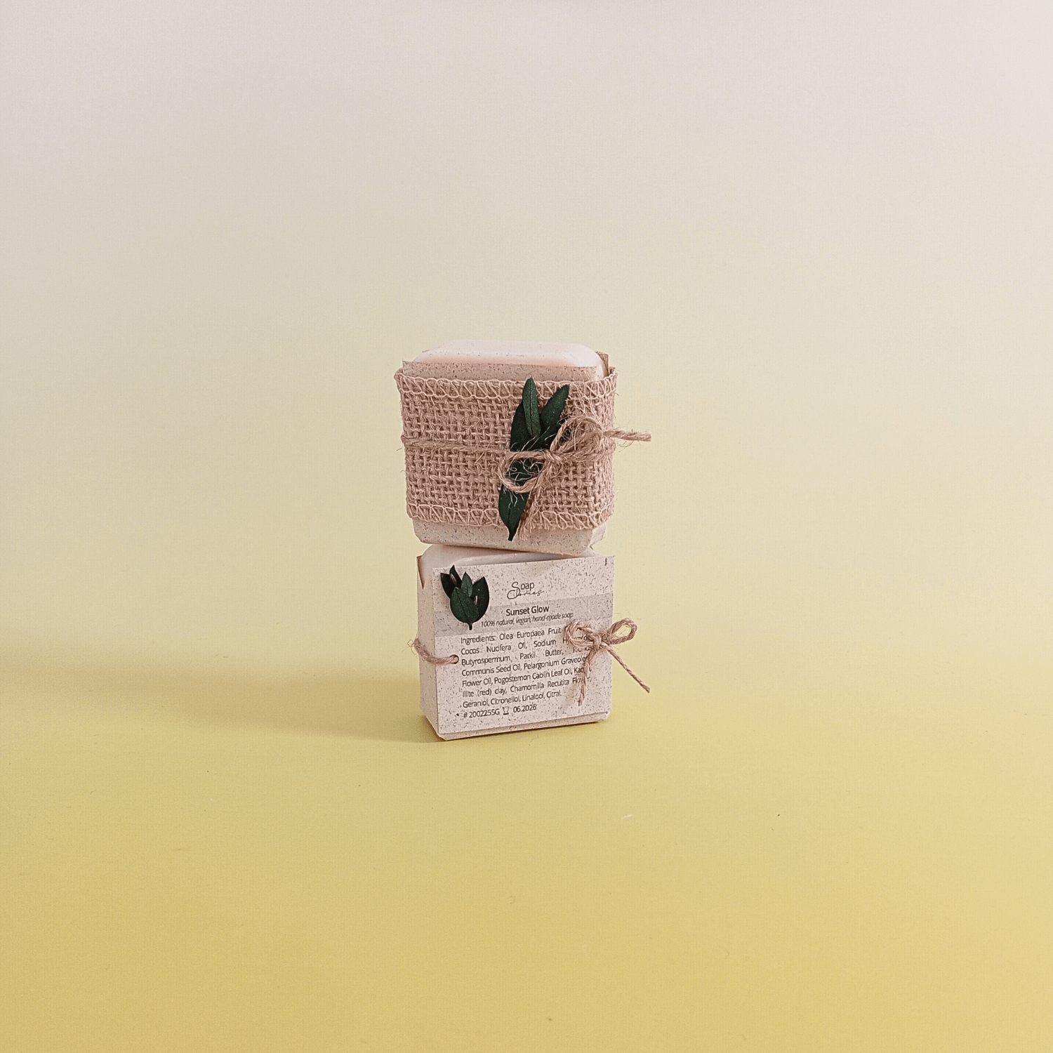 Natural soap bars wrapped in rustic burlap and twine with green botanical accent, displayed vertically with visible ingredients label on a soft yellow background.
