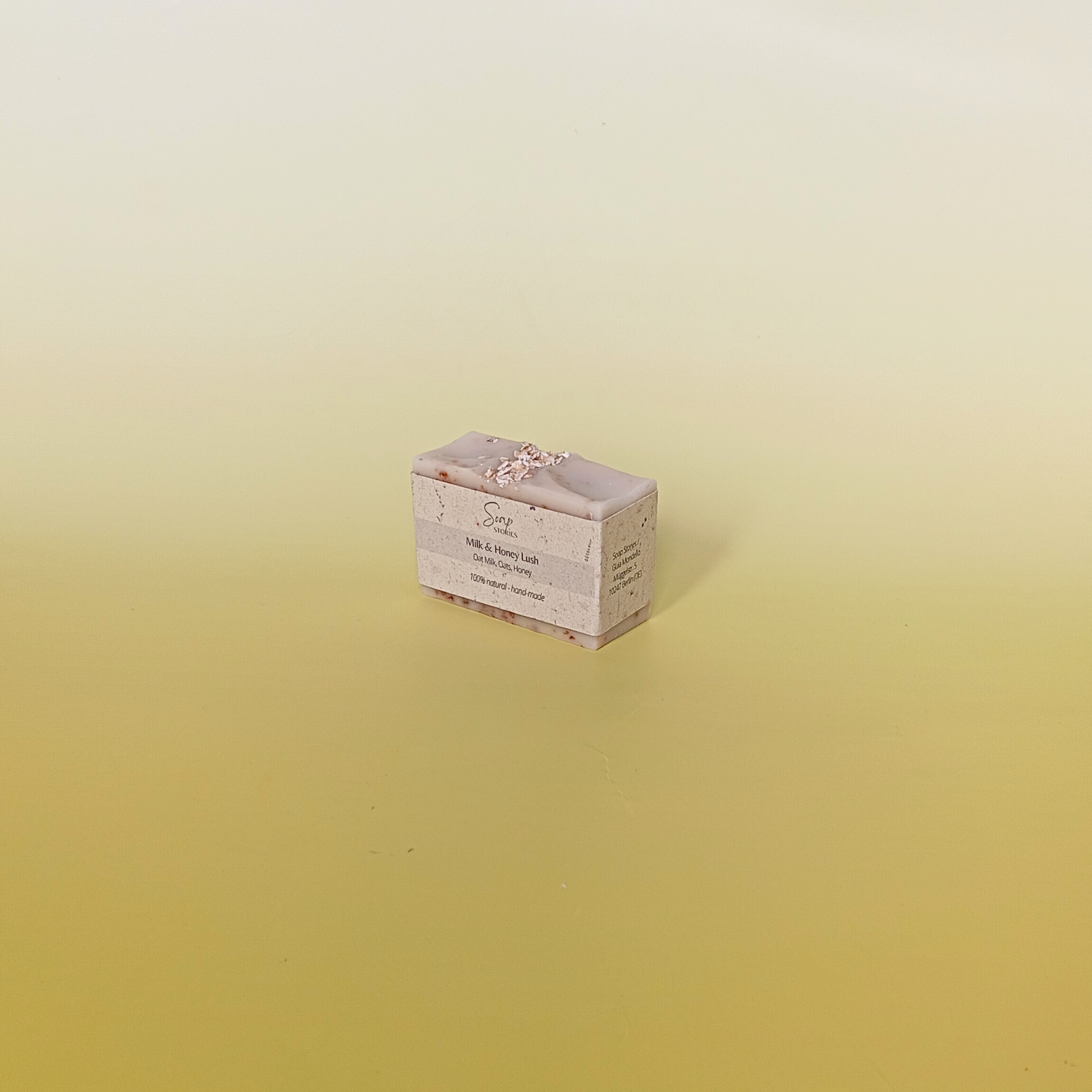 Handmade natural soap bar with a soft beige tone and visible oats, unscented and crafted with milk, honey, and oats, displayed on a pale yellow gradient background wrapped in grass paper sleeve