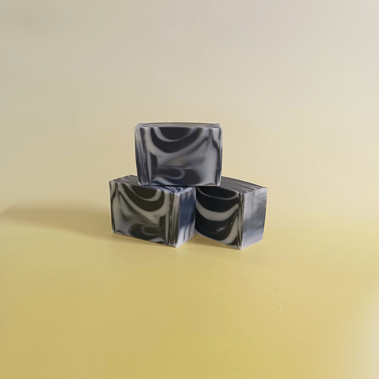 Three handmade natural soap bars with black and white swirl design, stacked together on a soft yellow background. Crafted with charcoal powder.