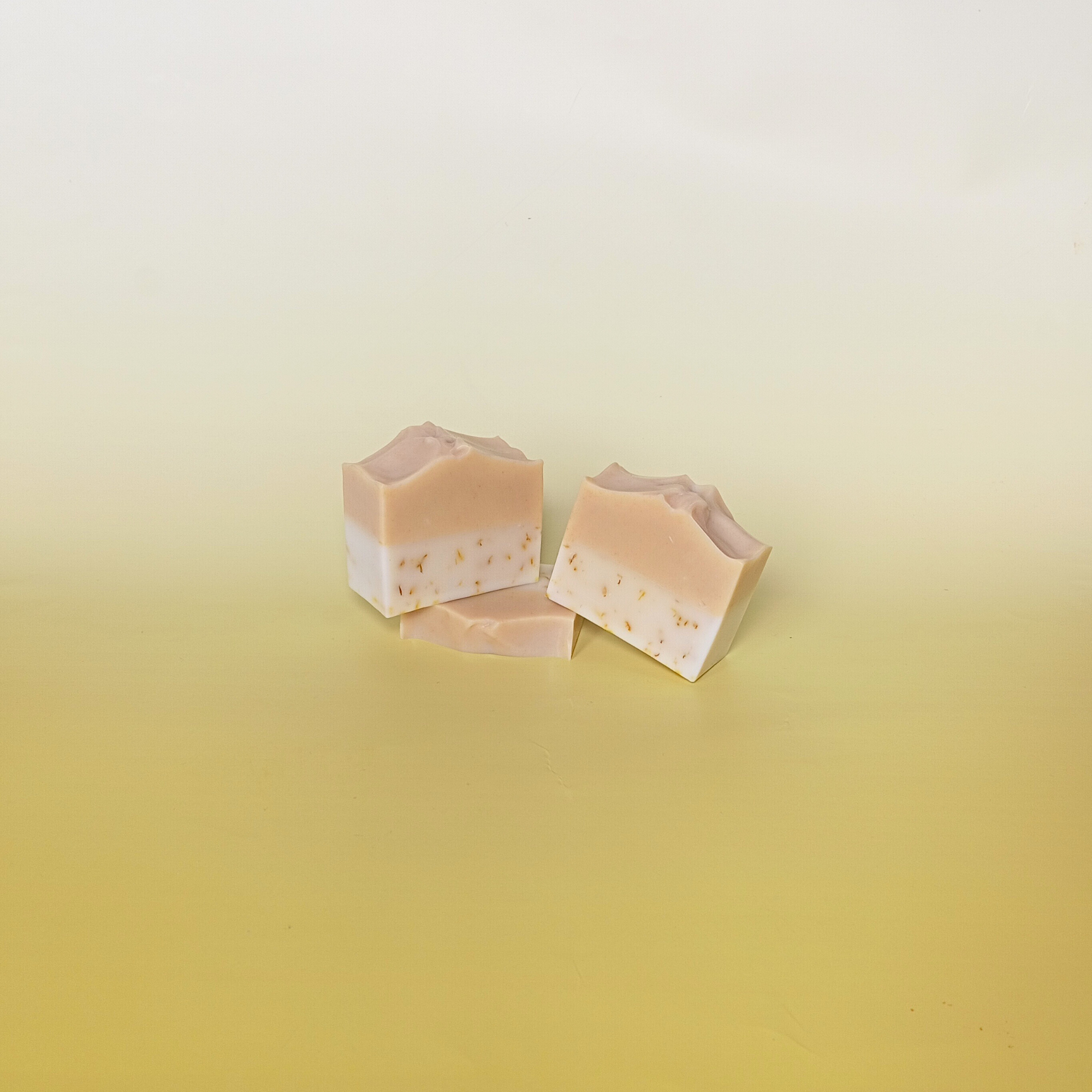 Three handmade two-tone natural soap bars with blush and cream layers, artistically stacked on a pale yellow gradient background, reflecting a soft and minimal design.