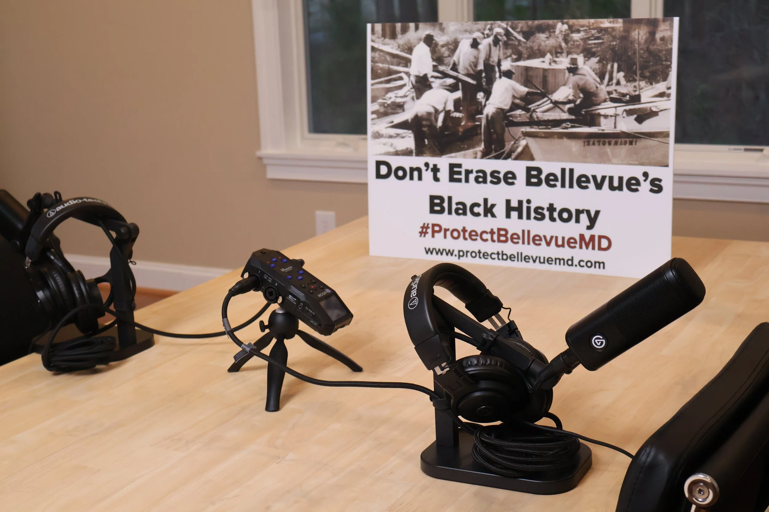 A conference table with two headsets, a sound mixer, and a sign that reads 'Don't Erase Bellevue's Black History' with hashtags #ProtectBellevueMD and a website address. The background shows a window and beige walls.