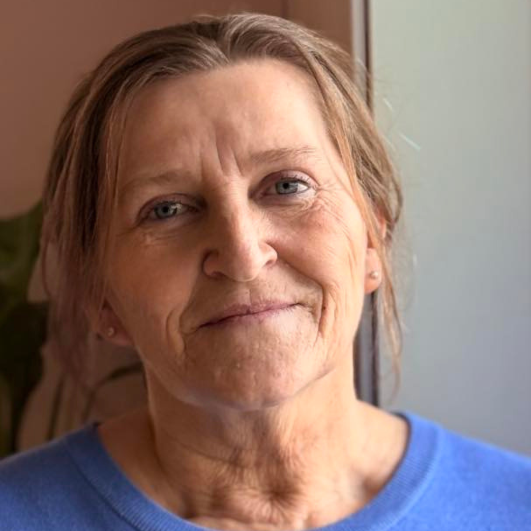 Et nærbilde av en eldre kvinne med kort, lysbrunt hår og lys hud, som smiler svakt, iført en blå genser.