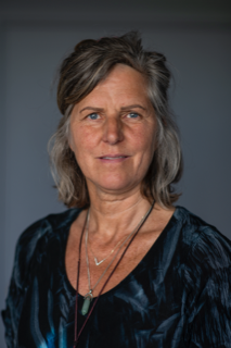 Bilde av en voksen dame med blå øyne, grålig hår og blå bluse. Hun har en krystall hengende rundt halsen