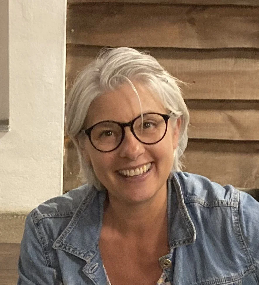 En kvinne med kort grått hår og briller smiler bredt, hun er iført en denimjakke.
