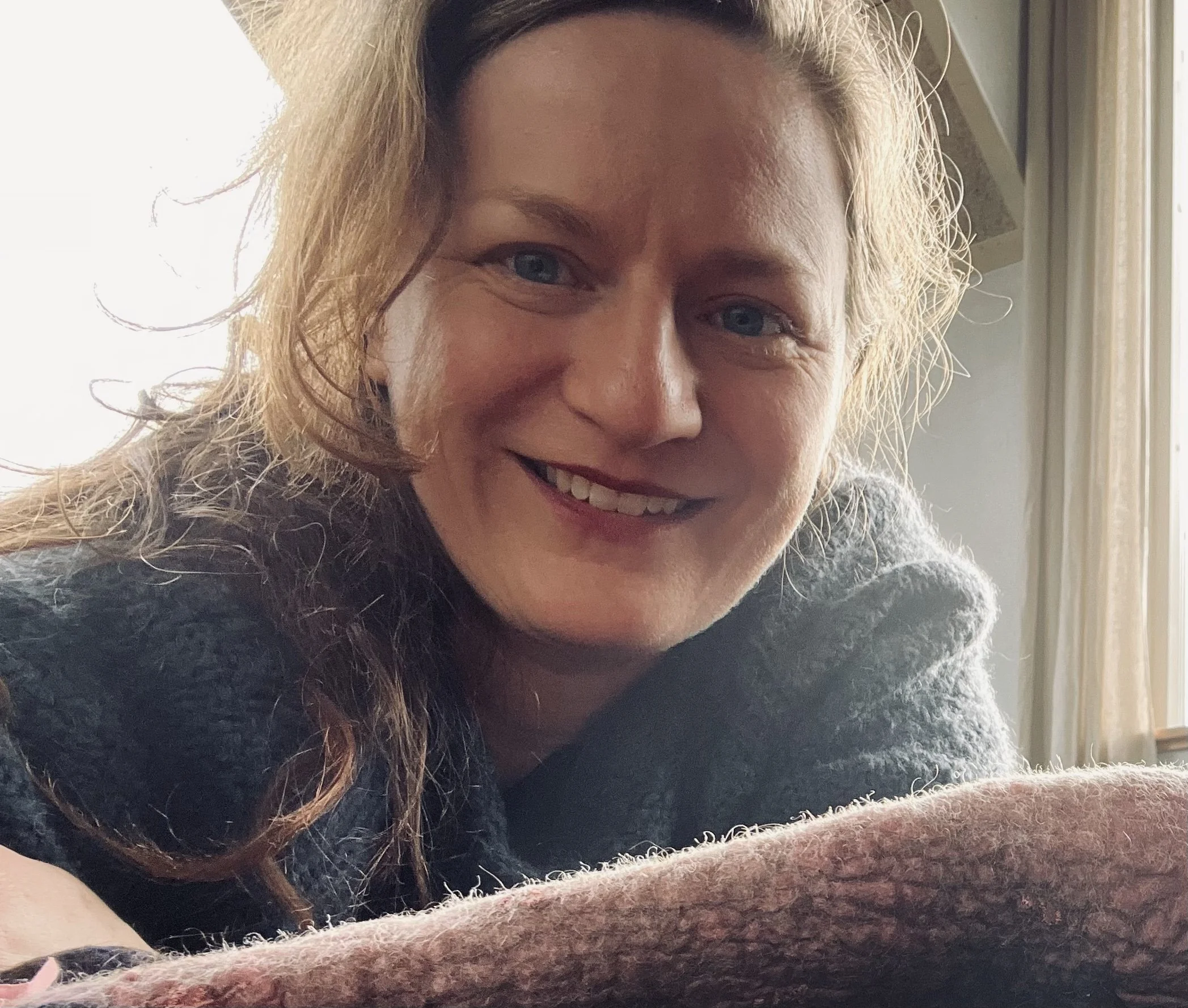 Et nærbilde av en person som smiler, med håret hengende over skuldrene og iført en mørk grå genser.
