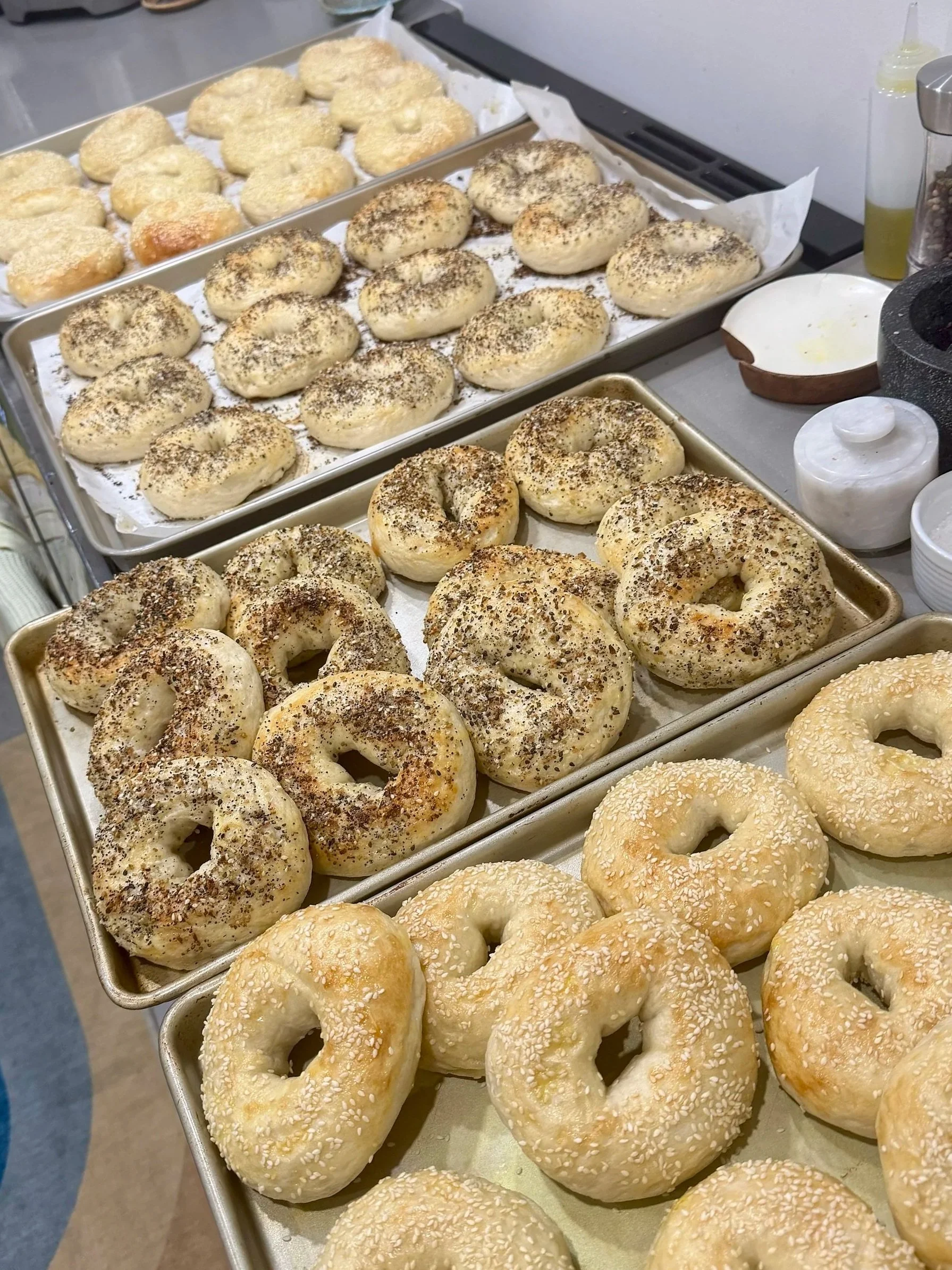 Baked bagels on trays with various toppings such as sesame, poppy seeds, and everything spice mix, ready for serving.