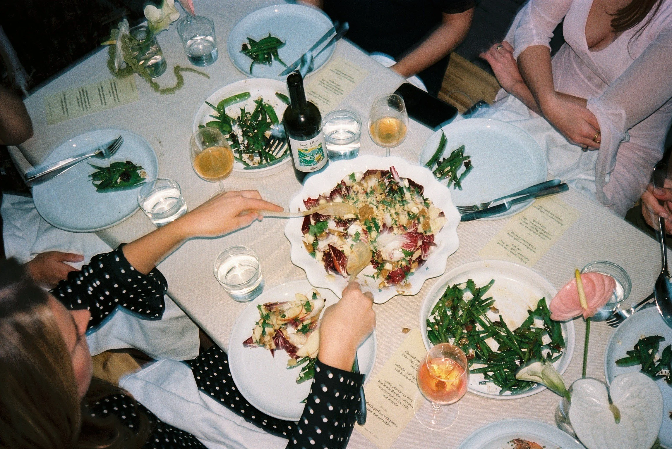 A table set with various dishes including a large salad, green beans, and drinks, with several people serving themselves and sitting around it.