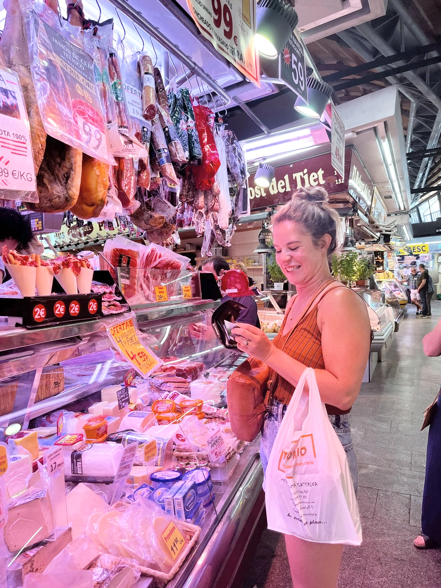 Britt shopping at a market's meat and cheese counter, smiling and pulling out money from her wallet while holding a shopping bag, with various meats and cheeses on display.