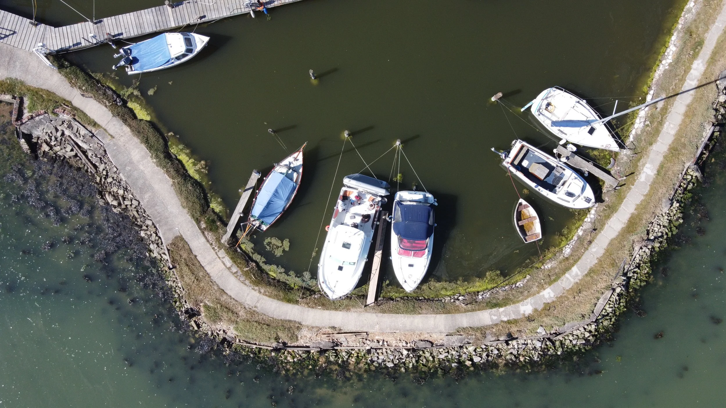 Aerial view of boats moored in a small marina with a dock and walking path, surrounded by green water and rocky shoreline.