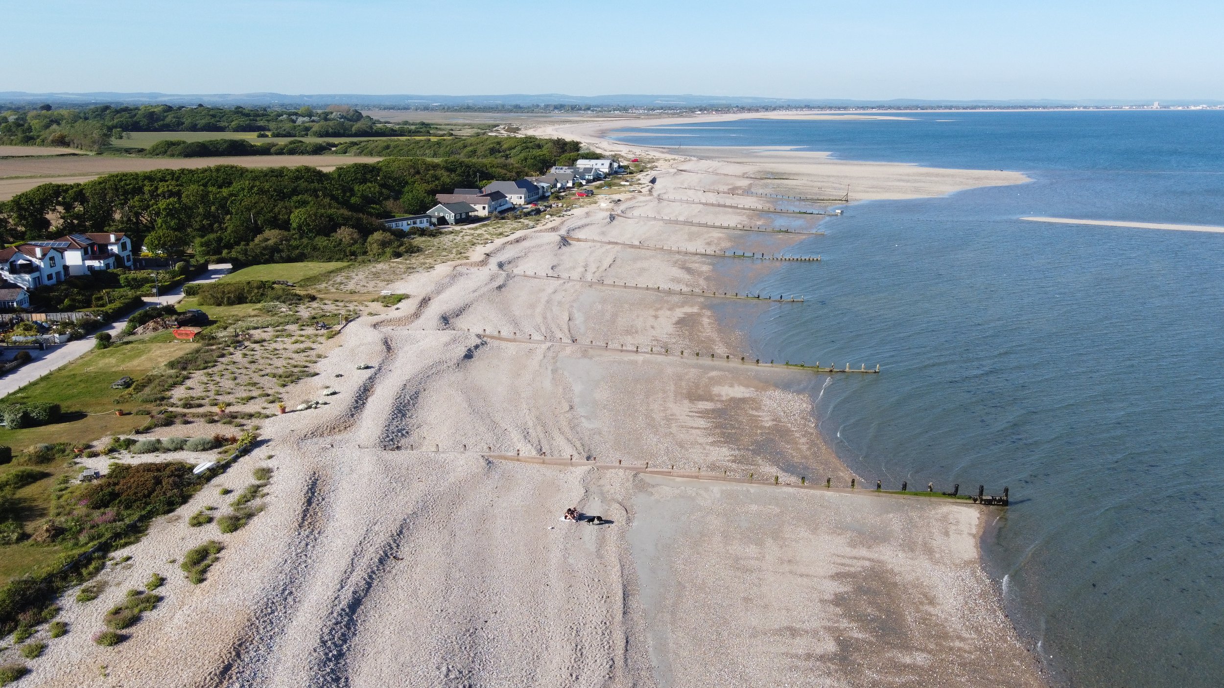 Aerial view of coastline with pebbled beach, groynes, houses, green vegetation, and blue sea.