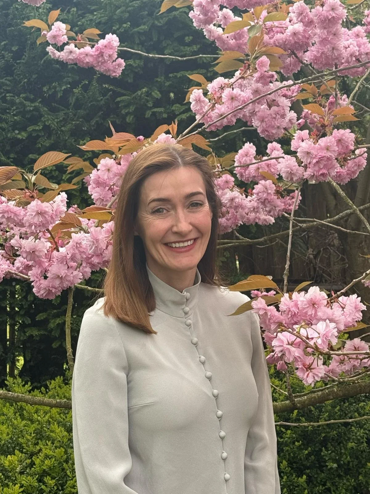 A smiling woman with shoulder-length brown hair standing in front of pink blossoming tree in a garden.
