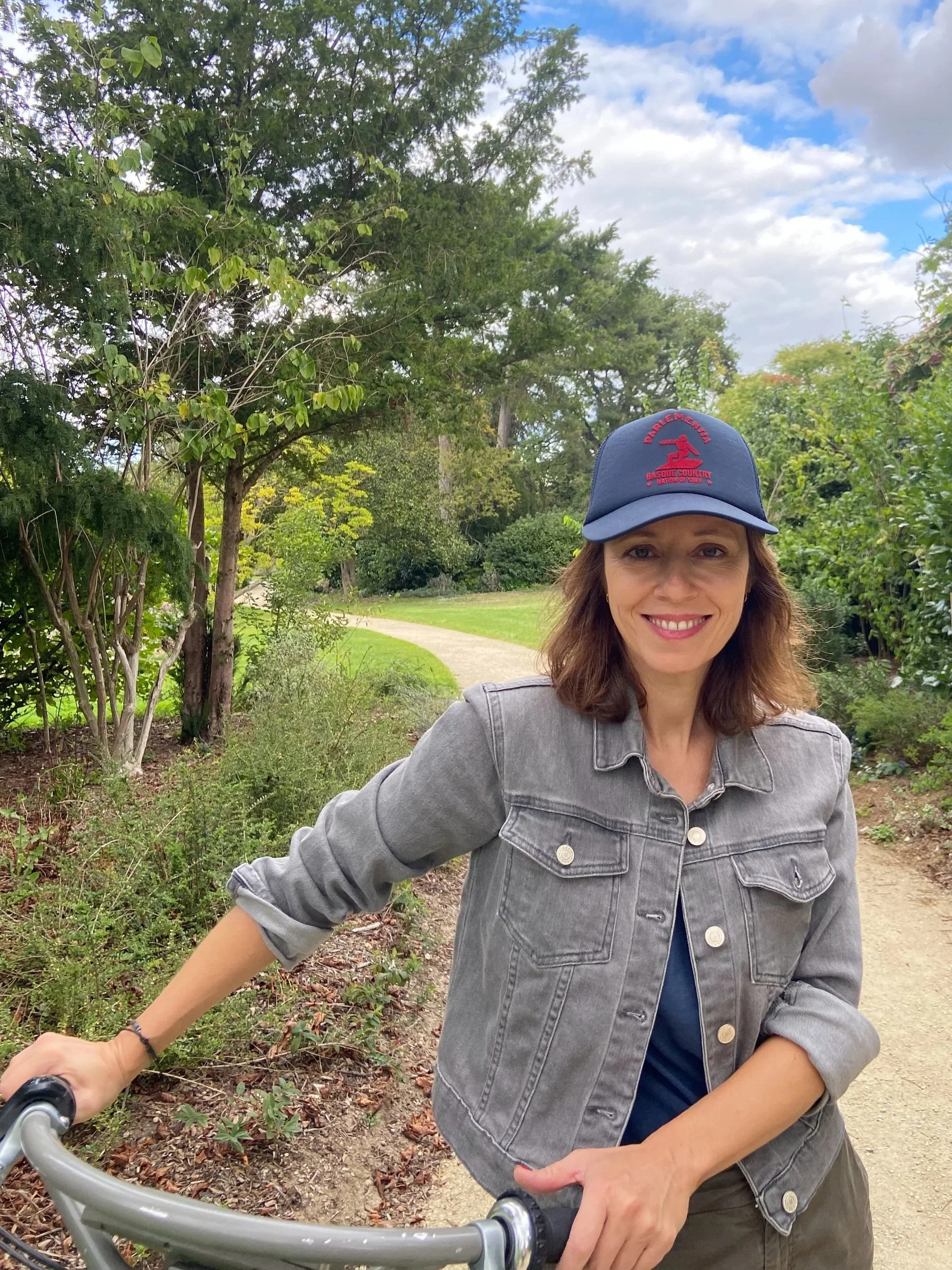 Une femme souriante portant une casquette et une veste en jean, tenant un vélo, en plein air dans un parc avec des arbres et un ciel partiellement nuageux.
