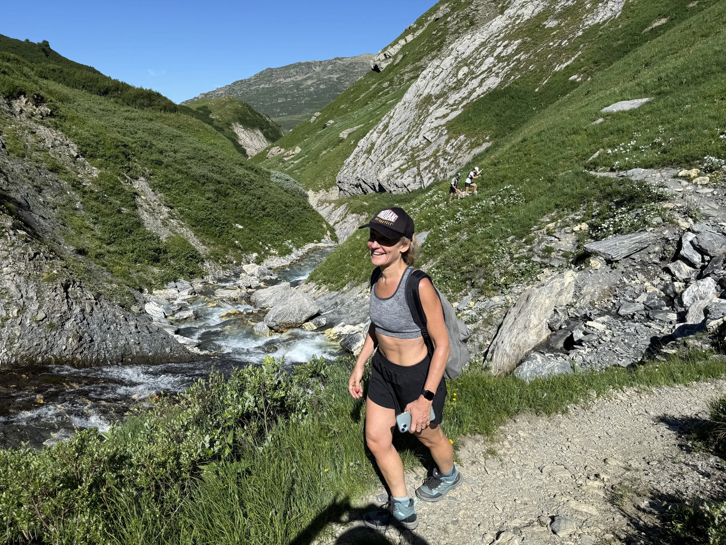 Femme en randonnée dans un paysage montagneux verdoyant avec un ruisseau et des montagnes en arrière-plan, portant un chapeau, un débardeur gris, un short noir et des chaussures de randonnée.