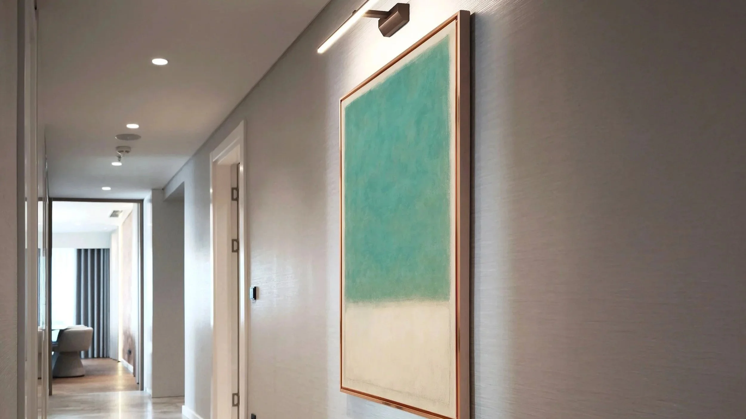 A hallway with beige walls and hardwood flooring, featuring a green and beige rug, with paintings and wall-mounted lights.