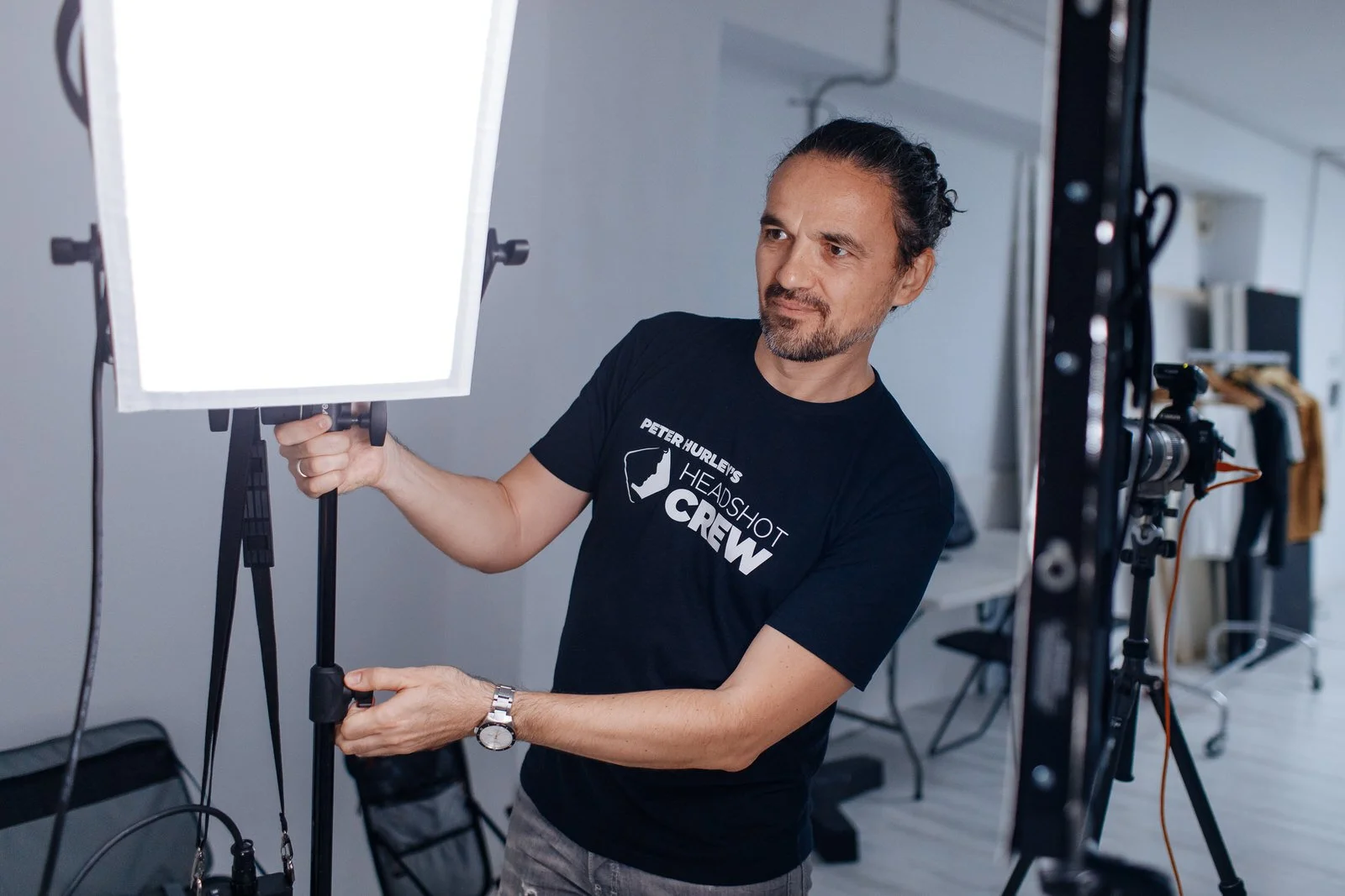 A man adjusting a lighting panel in a photography studio, wearing a black t-shirt that says 'Peter Hurley's Headshot Crew'.
