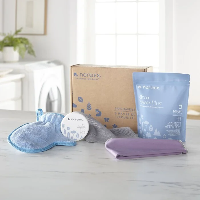 Cleaning supplies, including a brown box, a package of Norwex Ultra Power Plus detergent, a round container of Norwex cleaning paste, a gray microfiber cloth, a purple cloth, and a blue microfiber polishing cloth on a white marble countertop in a laundry room.
