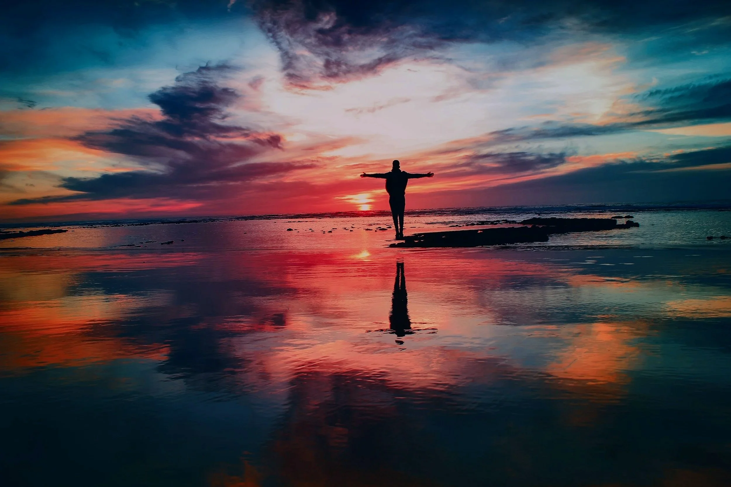 Person standing by the ocean at sunset with arms open, symbolising emotional release and healing through the Emotion Code.
