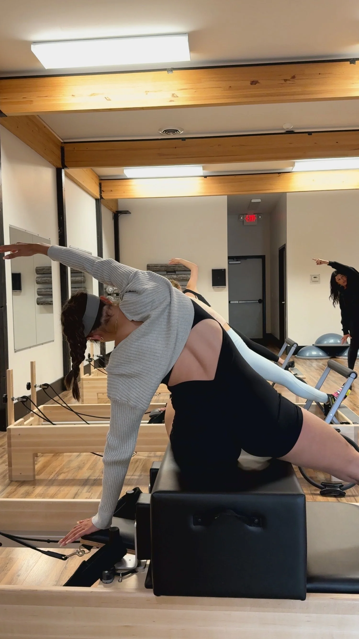 A woman in a Pilates studio doing a stretching exercise on her side, with other participants visible in the background.