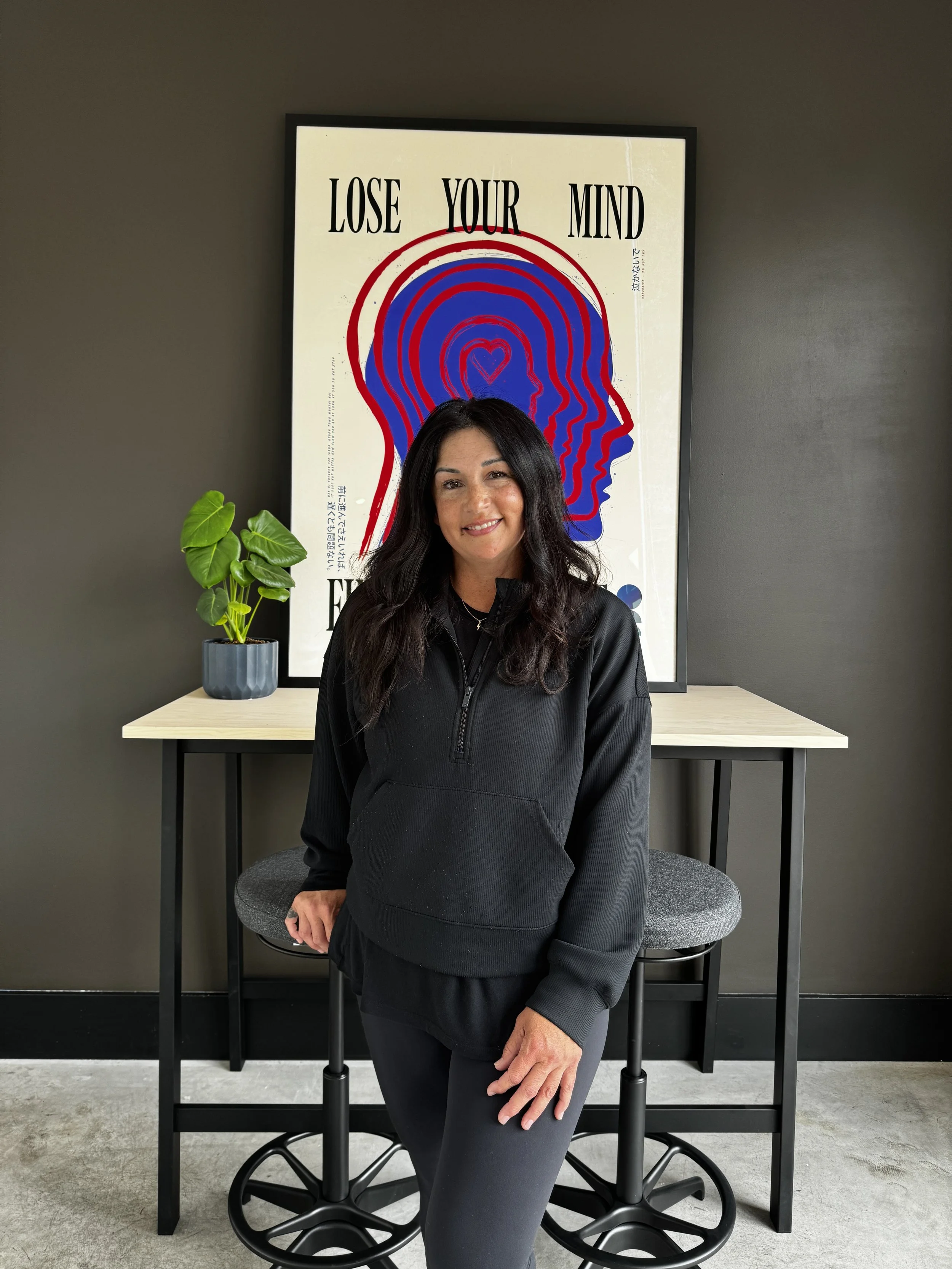 A woman with long black hair smiling, standing in front of a framed poster that says 'Lose Your Mind' with a graphic of overlapping blue and red outlines of a human head and a heart, with a small plant on her left and a light-colored table behind her.