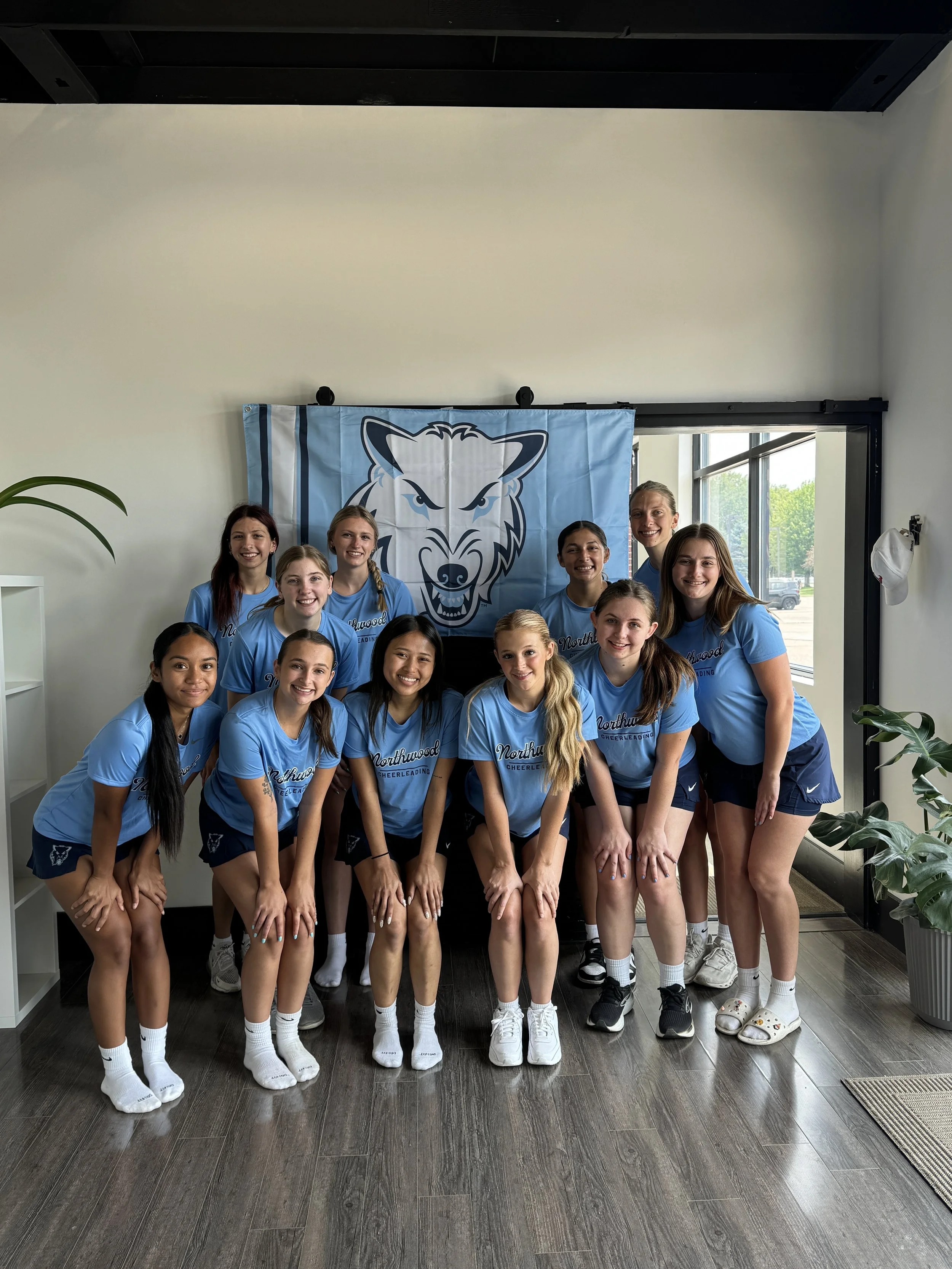 A group of eleven cheerleaders dressed in light blue shirts and navy shorts, posing indoors in front of a Northwood Cheerleading banner with a wolf logo.