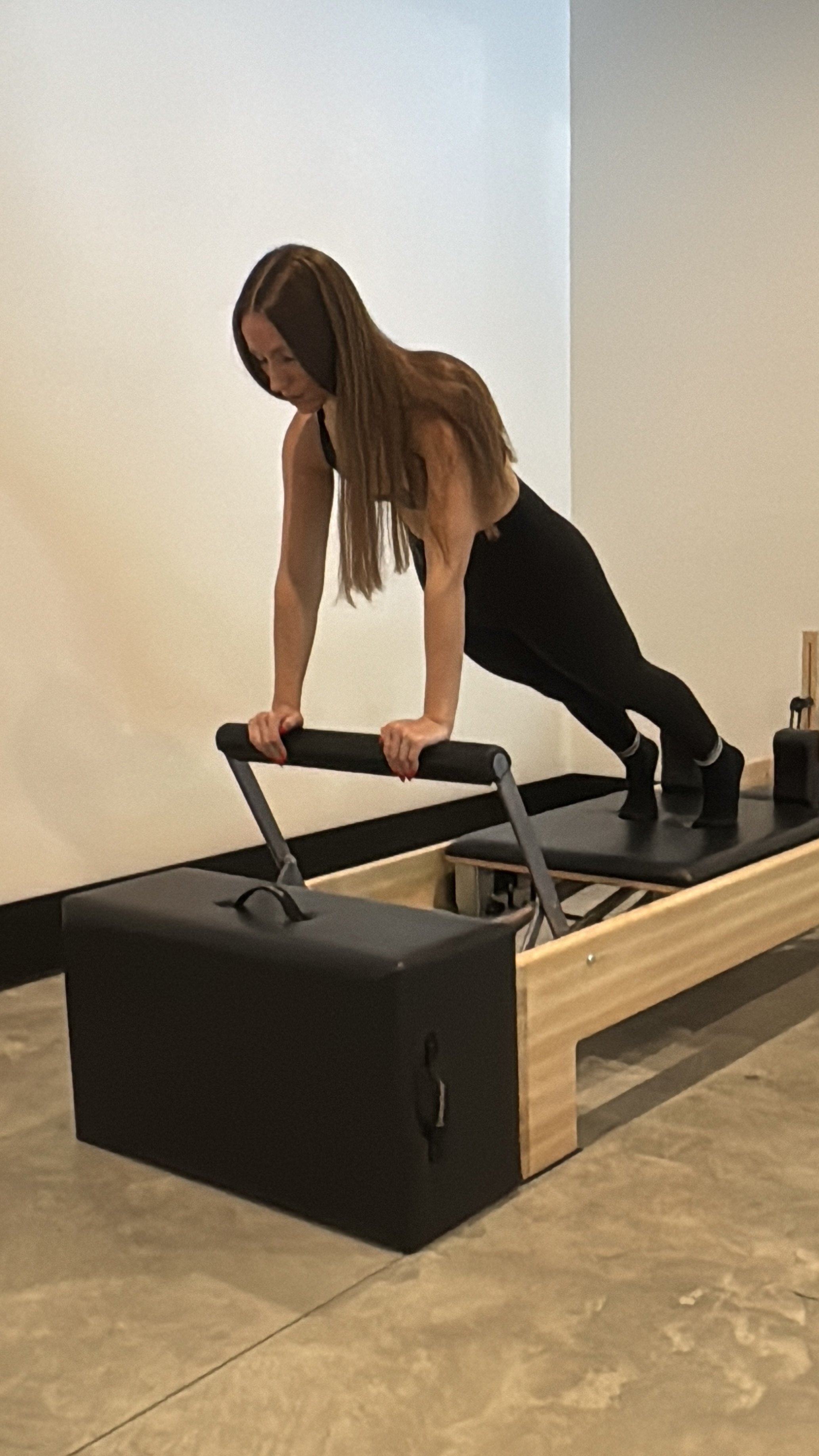 A woman doing a plank exercise on a Pilates reformer machine with a black box at the end.