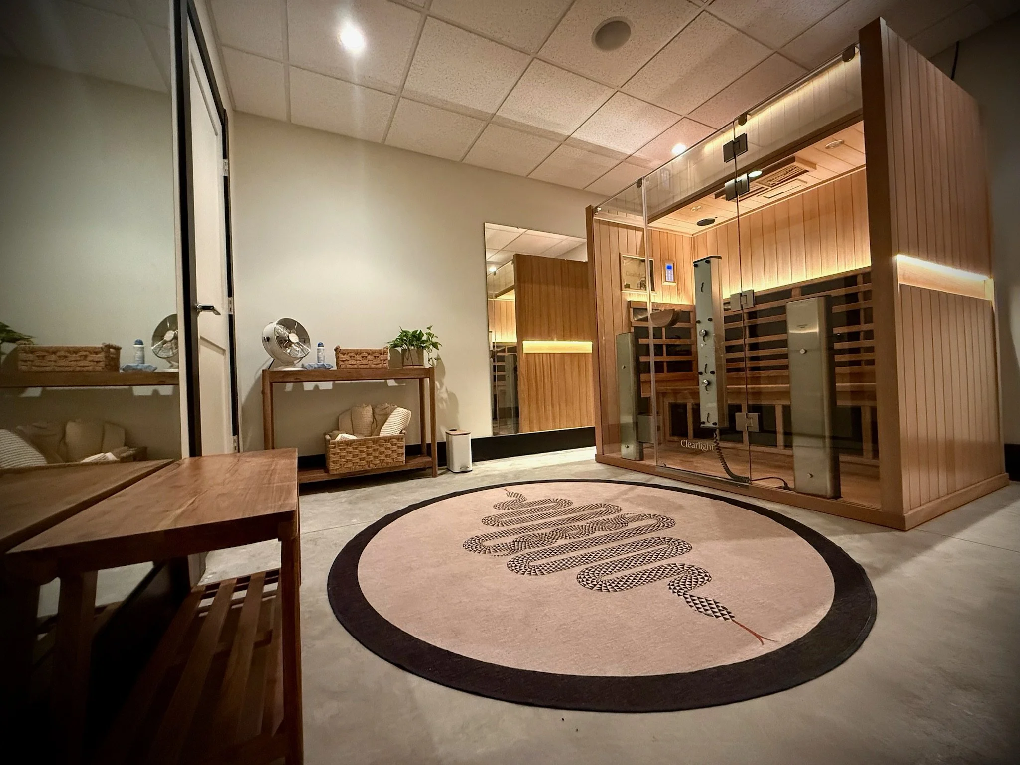 A modern infrared sauna room with glass walls, wooden interior, a bench, and a control panel. The room has soft lighting and a circular rug with a snake design.