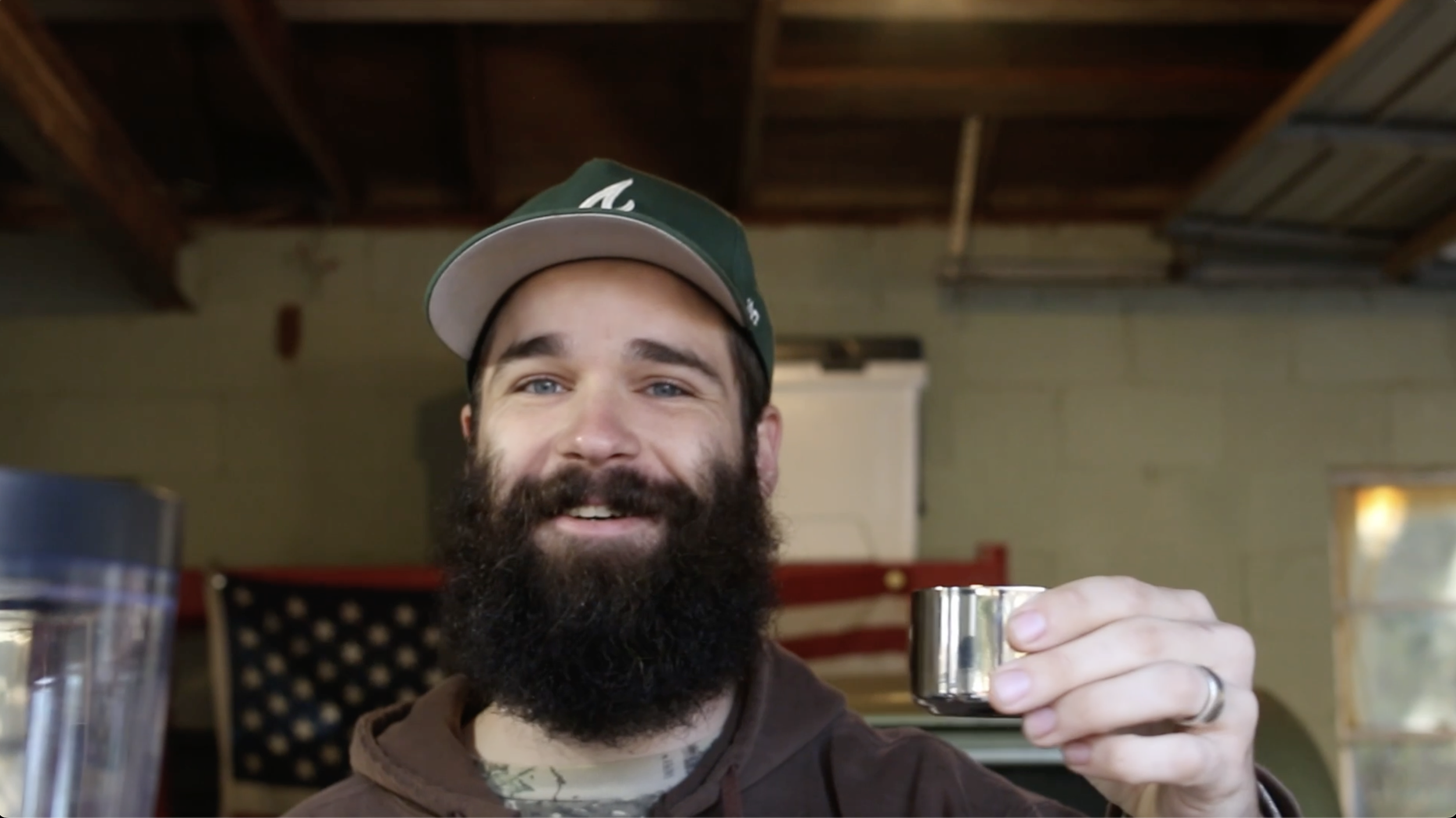 A man with a beard and mustache wearing a green cap smiling and holding a shot glass. Indoor setting with a bright window and an American flag in the background.