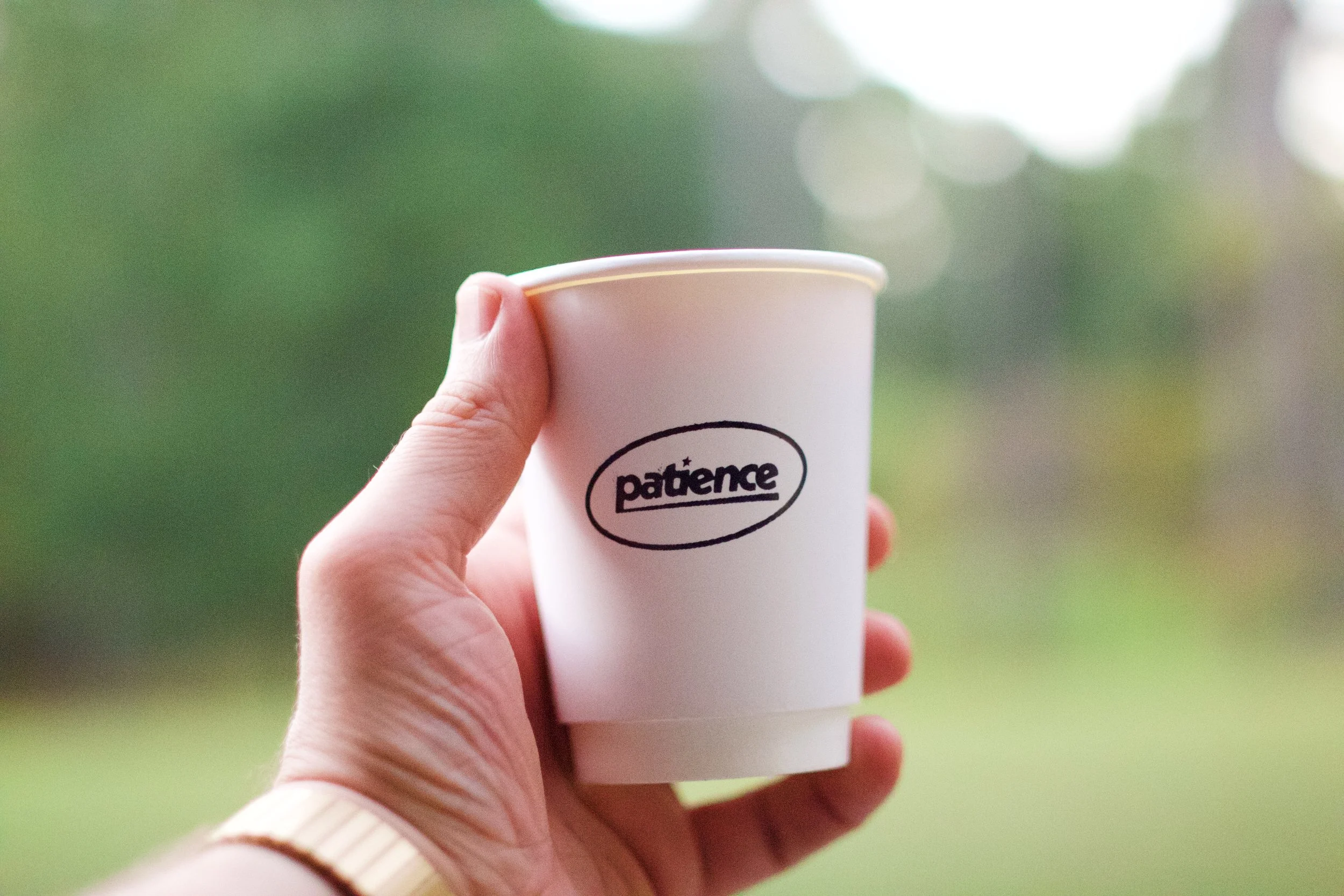 A hand holding a white disposable cup with the word 'patience' written on it, outdoors with a blurry green background.