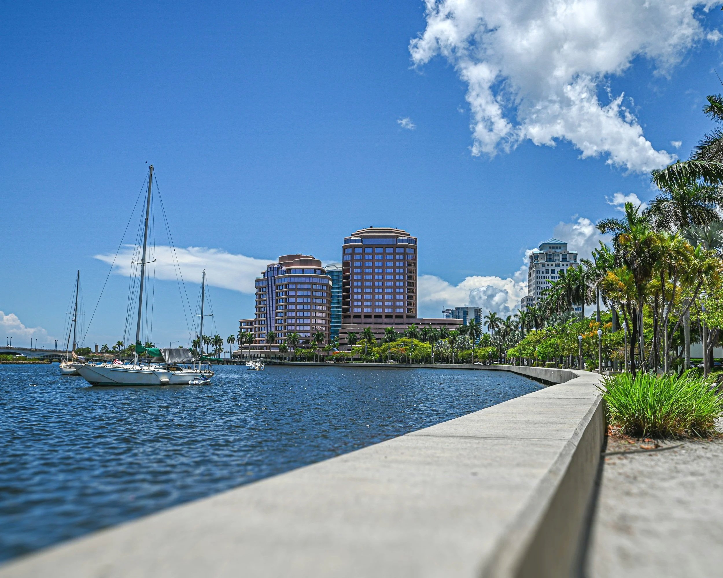 View of sailboats docked at a marina, with tall modern buildings and palm trees in the background under a partly cloudy blue sky.