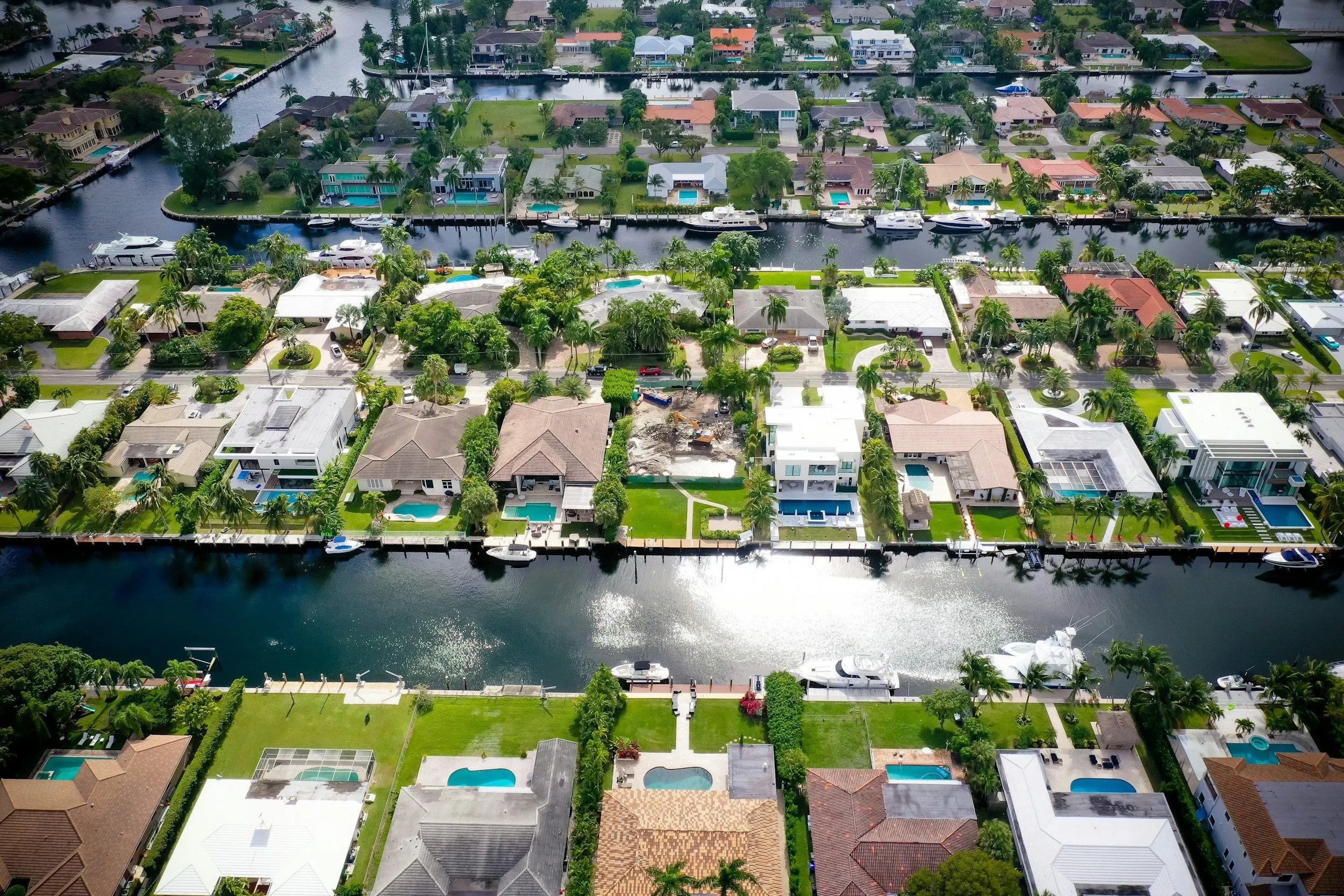 Aerial view of a residential neighborhood with houses, swimming pools, palm trees, and a canal with boats.