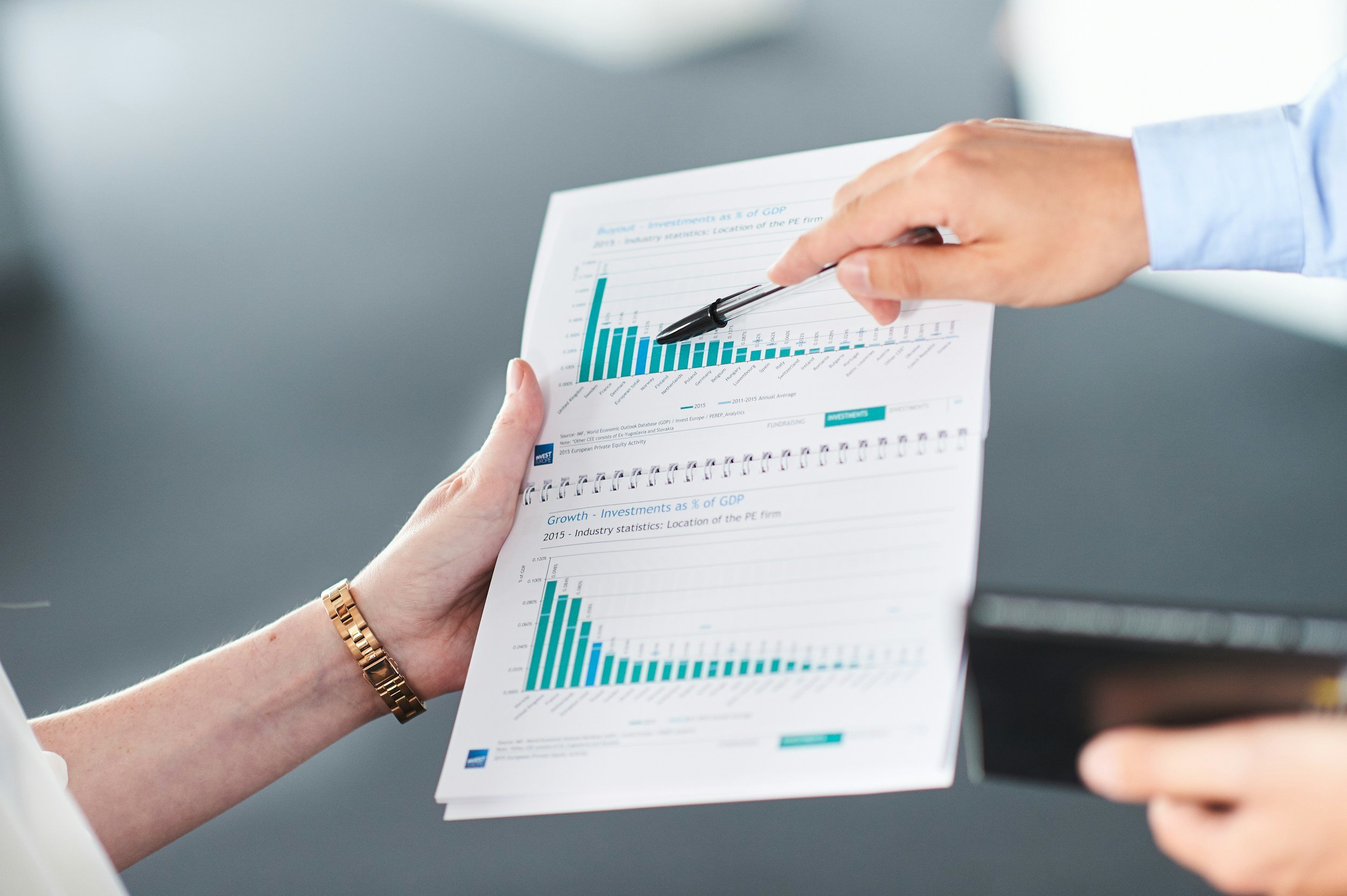 A person with a gold watch and a light blue shirt holding a sheet of paper with bar graphs and data charts. The person's hand points with a pen on the paper, which appears to contain financial or investment information.