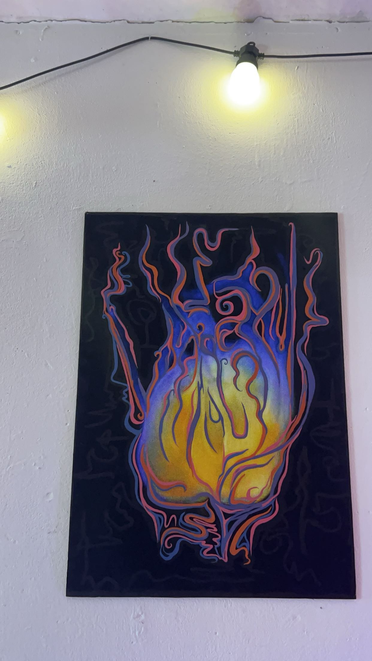 Abstract painting with swirling lines and colors resembling blue and yellow flames on a black background, hanging on a textured wall under a light bulb.