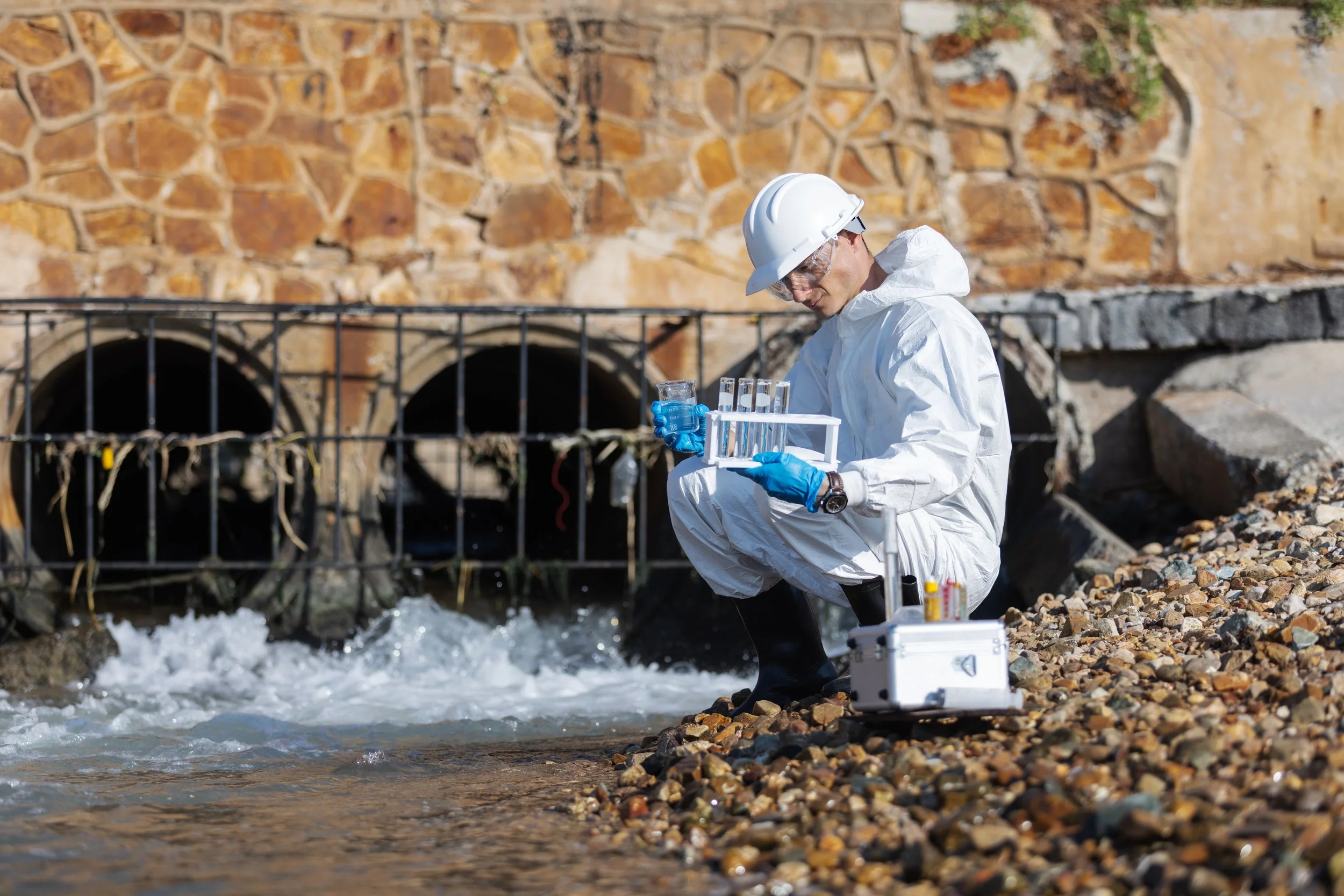 Environmental scientist in protective gear collecting water samples by a river with a stone wall and drainage pipes in the background.
