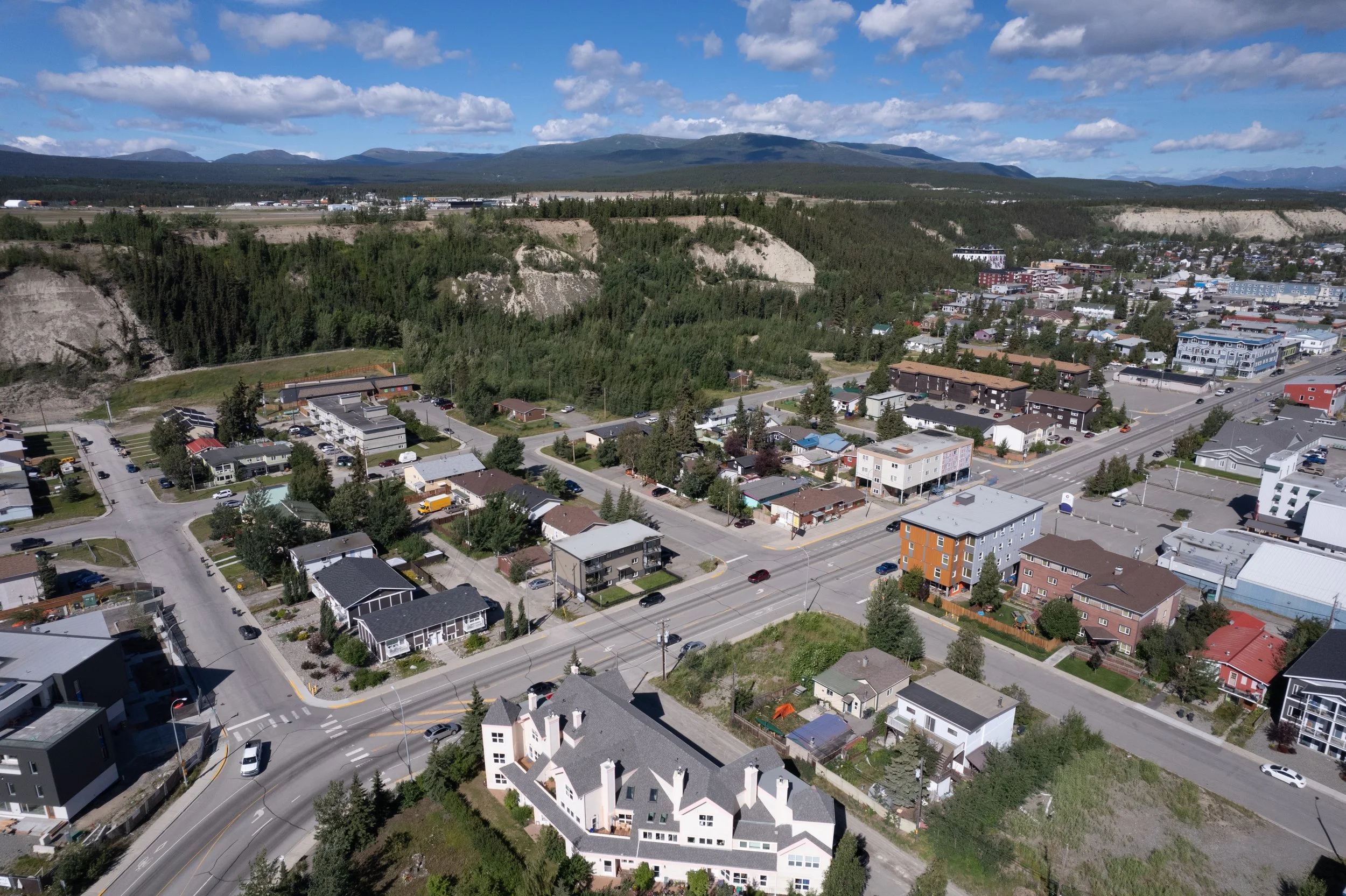 Aerial view of a small town with residential houses, commercial buildings, and streets surrounded by green hills and mountains under a partly cloudy sky.