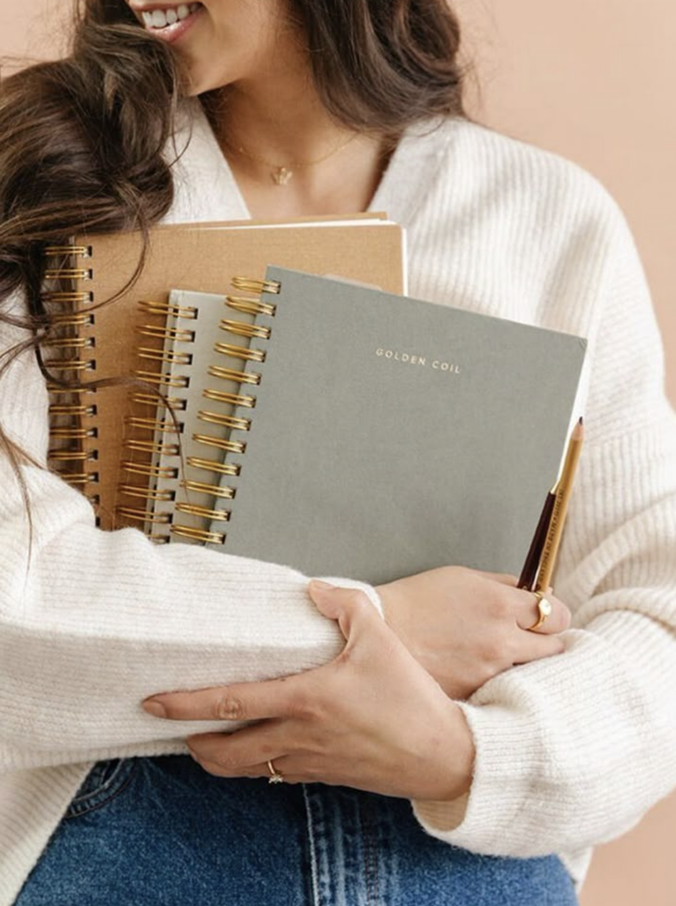 A woman holding three spiral-bound notebooks and a pen, all close to her chest. She is smiling and wearing a white sweater and blue jeans.