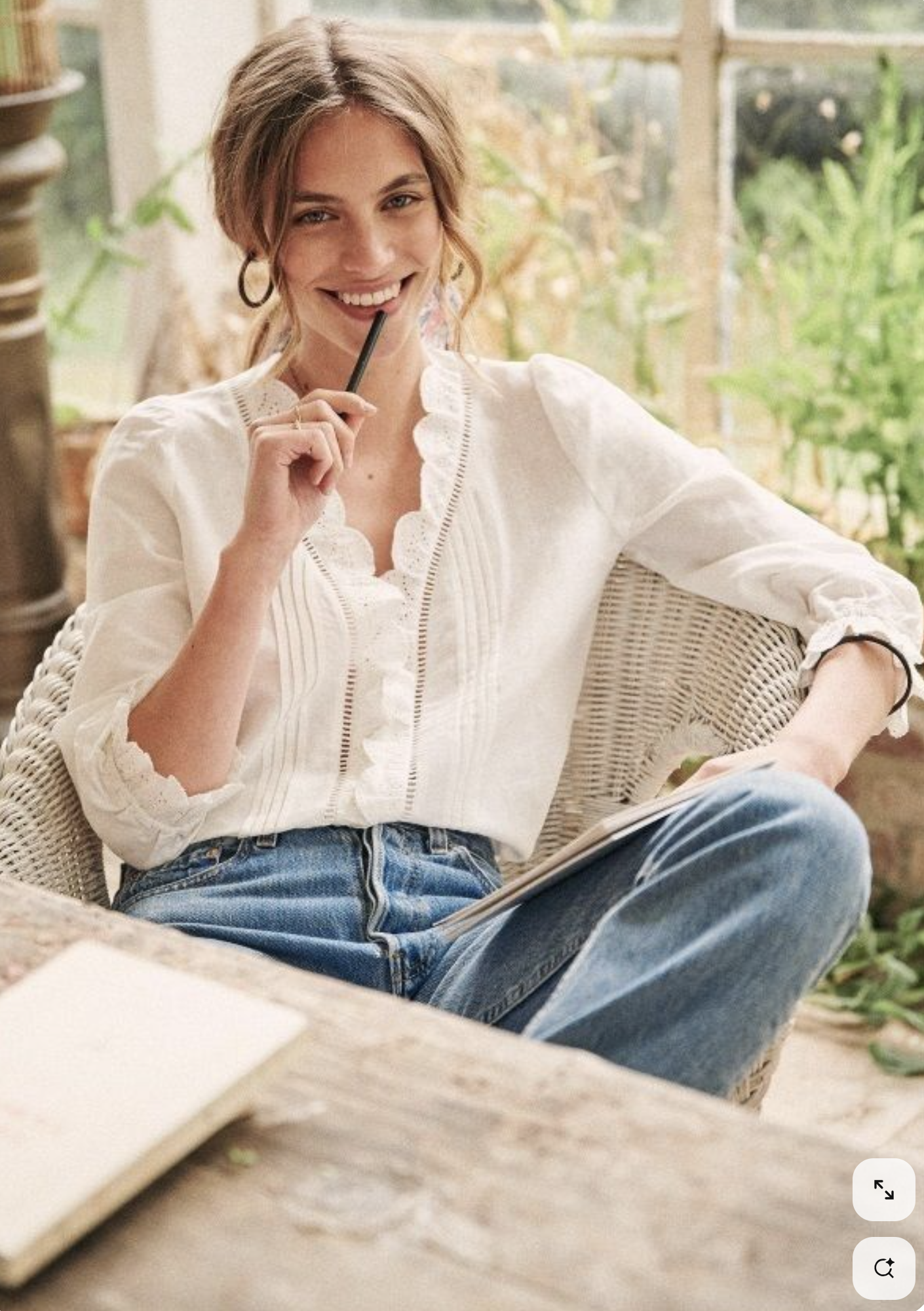 A woman sitting on a wicker chair in a well-lit, homey space, smiling and holding a pen near her face with a notebook on her lap, wearing a white blouse and blue jeans.
