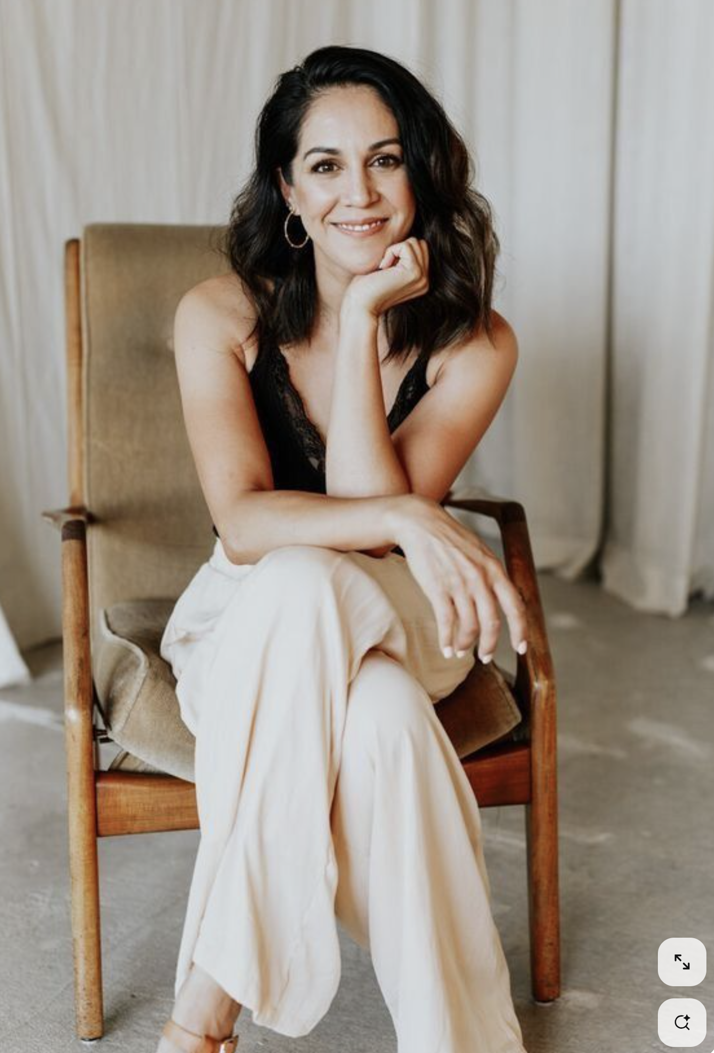 A woman sitting on a wooden chair with beige upholstery, wearing a black lace top and cream-colored pants, smiling at the camera with her hand resting on her chin.