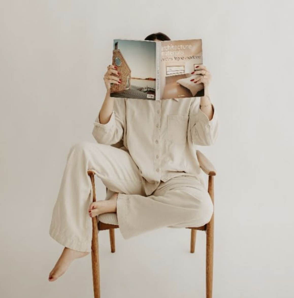 A person sitting cross-legged on a wooden chair, wearing beige pajamas, and holding a large magazine that covers their face. The background is plain white.