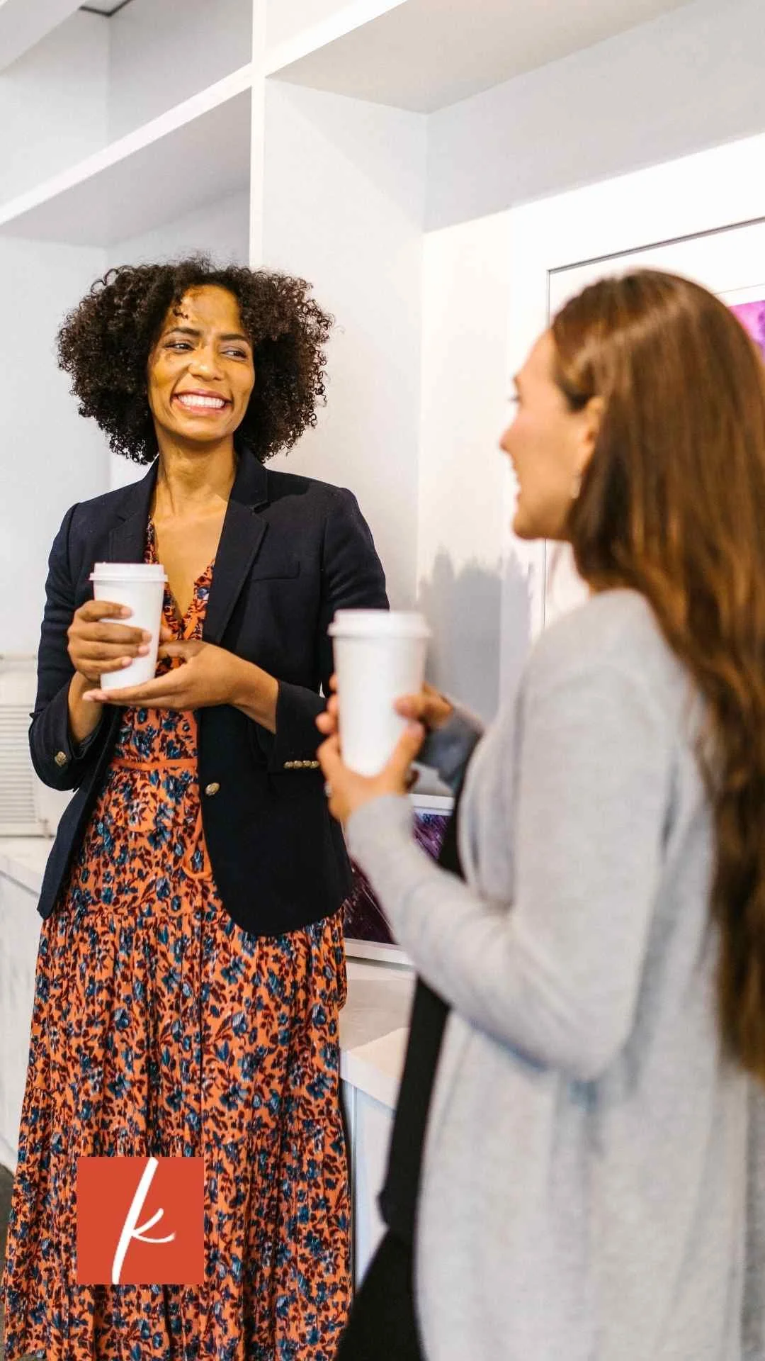 Two women smiling and talking while holding coffee cups in a bright, modern office space.